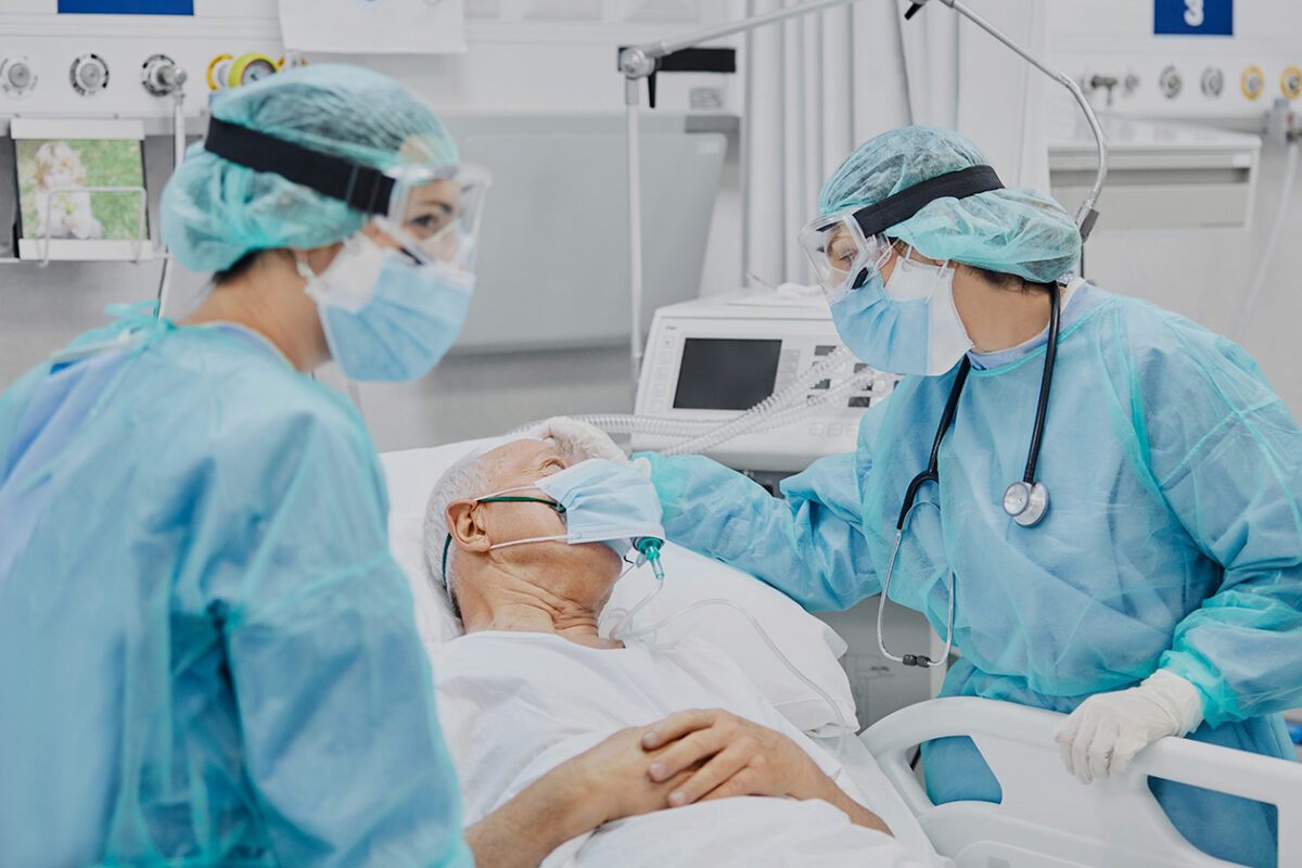 Photo shows an elderly man in a hospital bed with a breathing apparatus while two doctors in surgical gowns, masks and goggles care for him