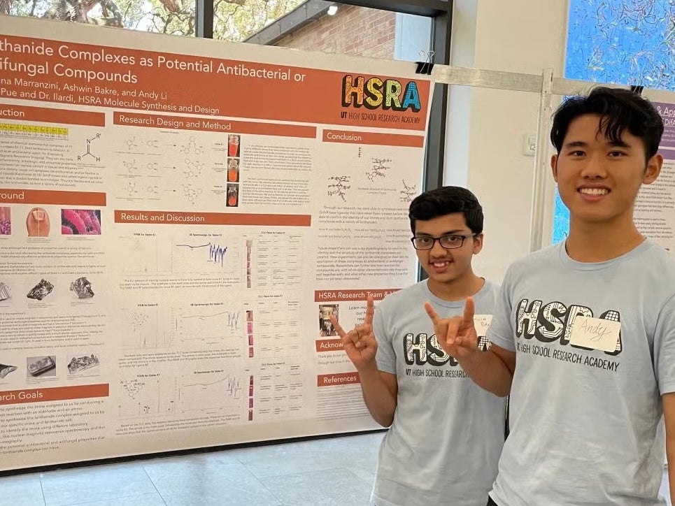 Two students in HRSA tshirts hold up the hook 'em horns hand sign and stand next to a research poster.