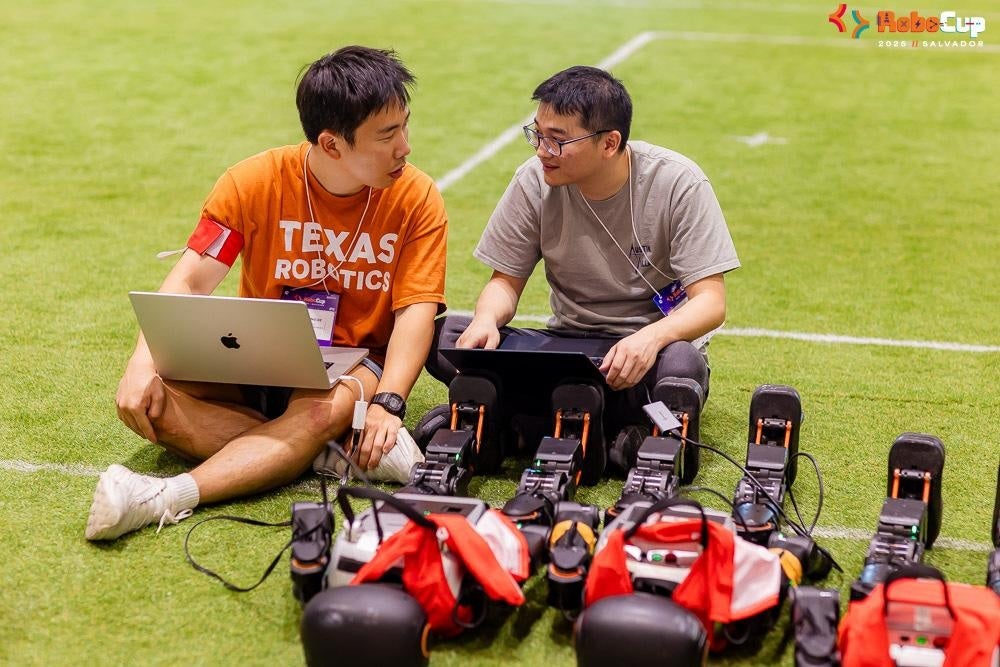 Young men with laptops, one wearing a Texas Robotics shirt, consult with one another while sitting on what looks like a soccer field in front of three humanoid robots.