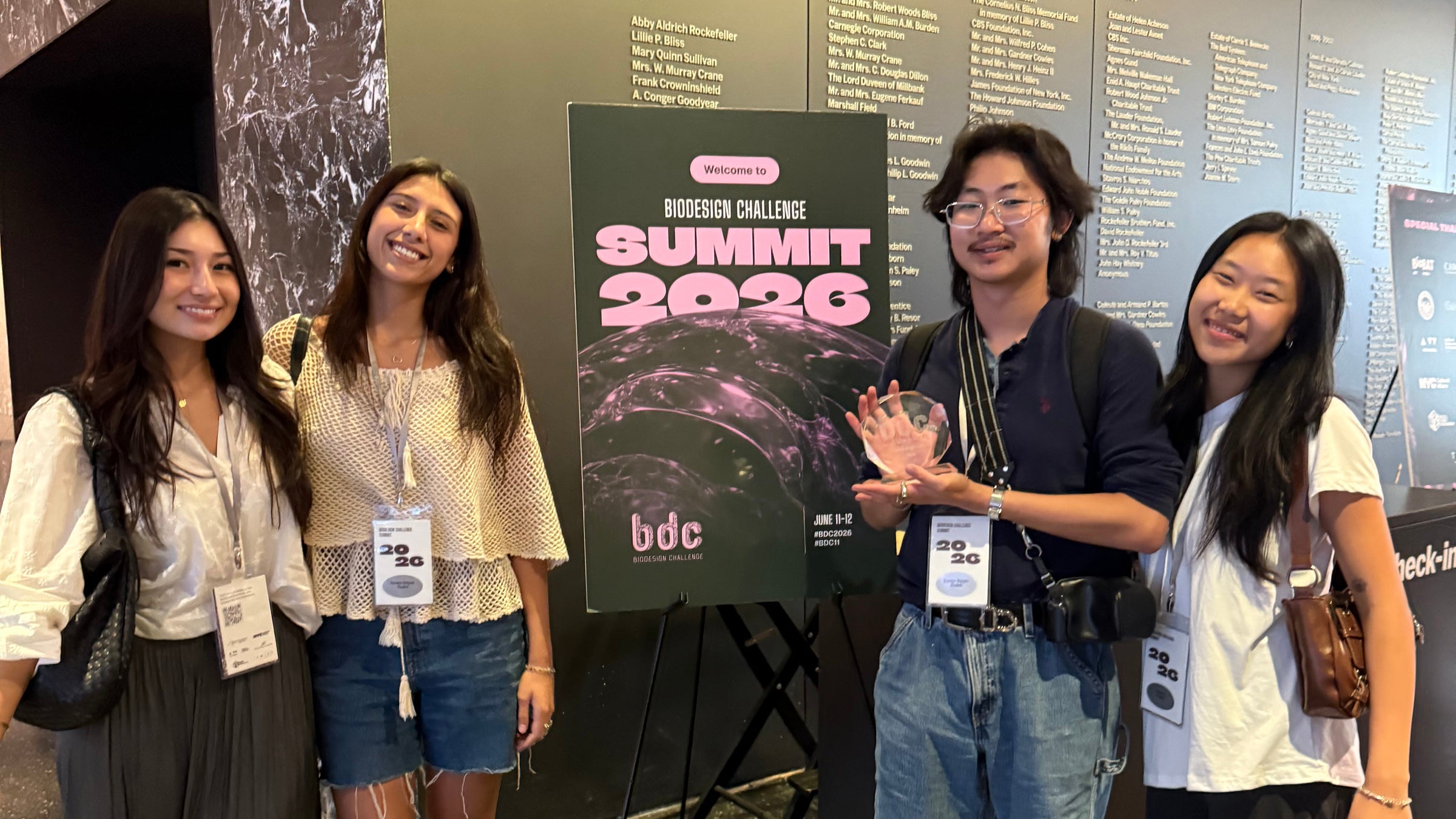 Four college students stand nest to a poster that reads Biodesign Challenge Summit 2026. One is holding a small glass trophy.