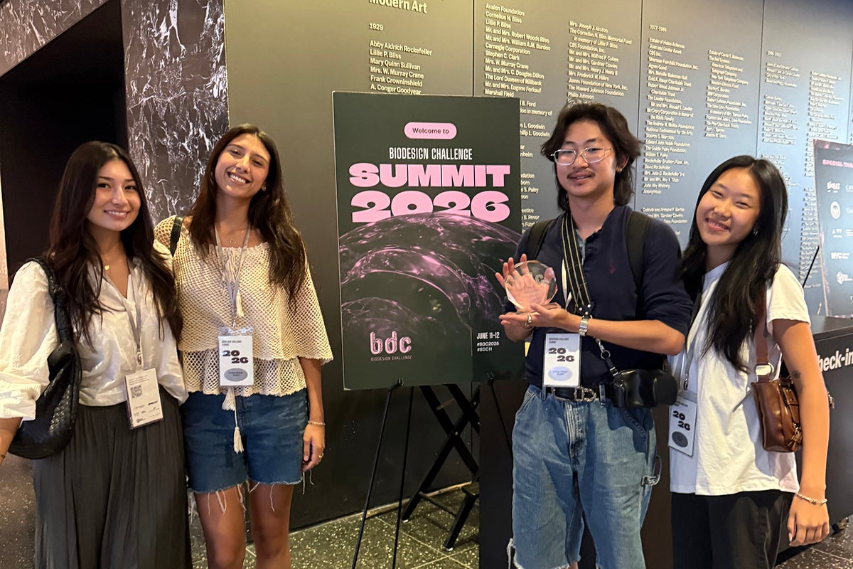 Four college students stand nest to a poster that reads Biodesign Challenge Summit 2026. One is holding a small glass trophy.