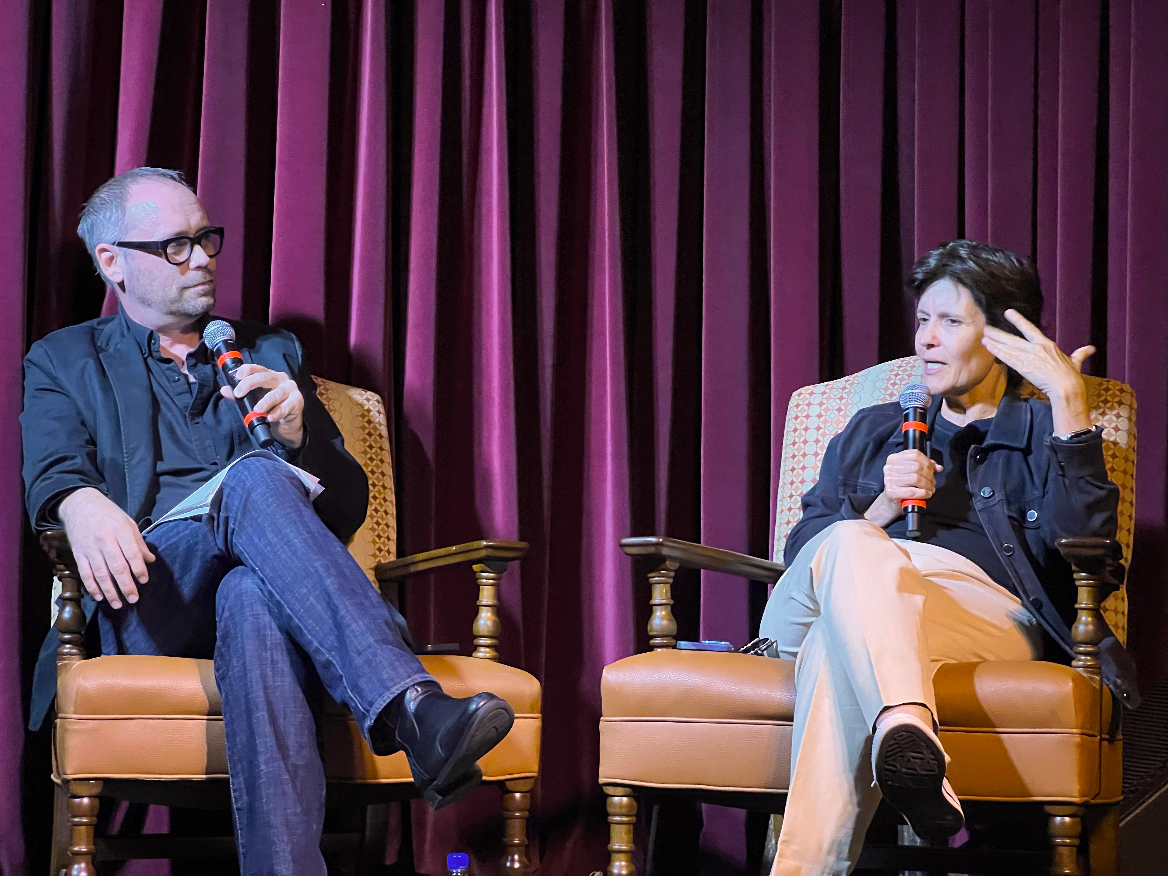 A man and a woman sit in armchairs on a stage and speak into handheld microphones
