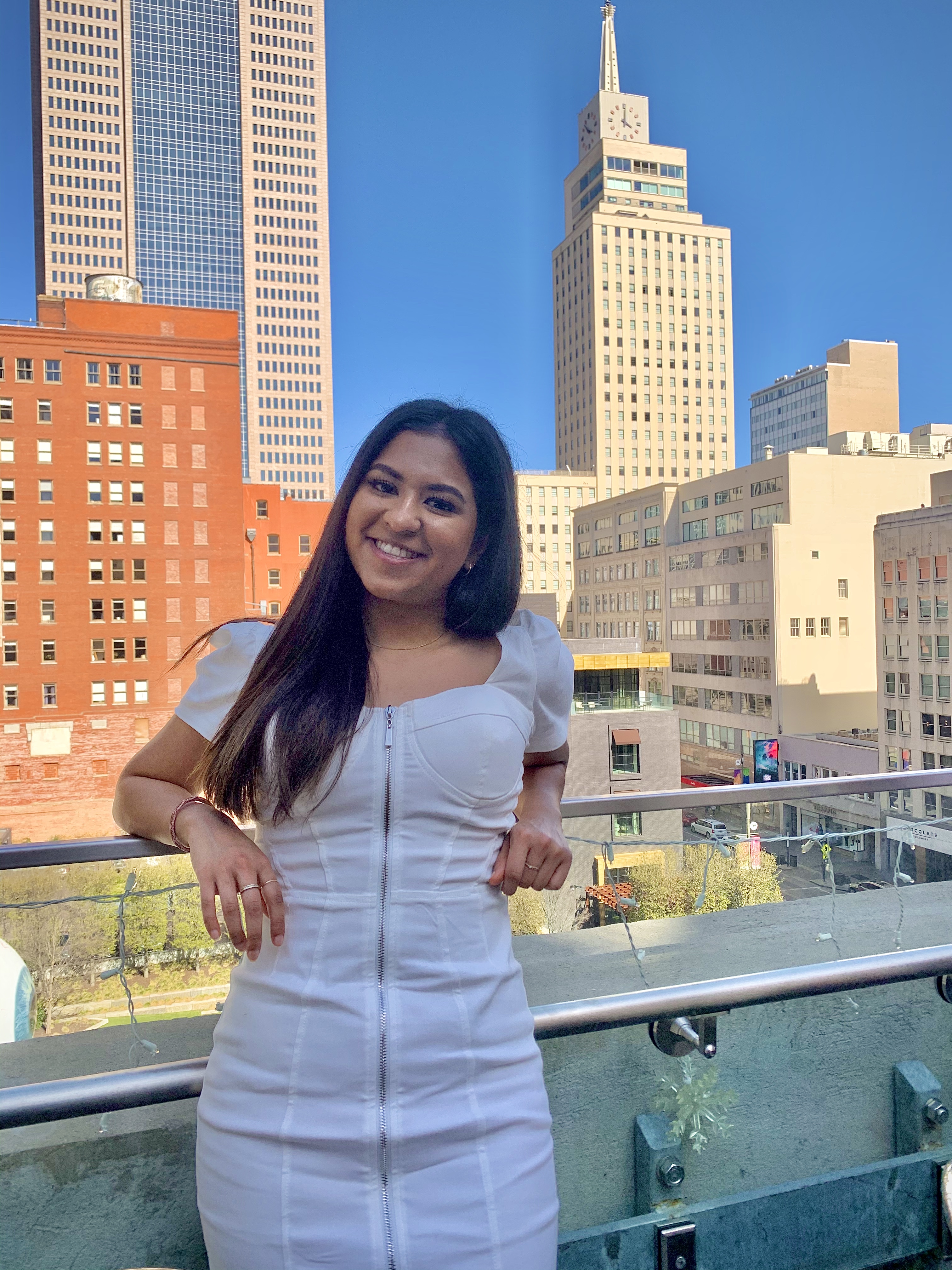 A young woman in a white dress stands on a balcony smiling with a city 's buildings behind her