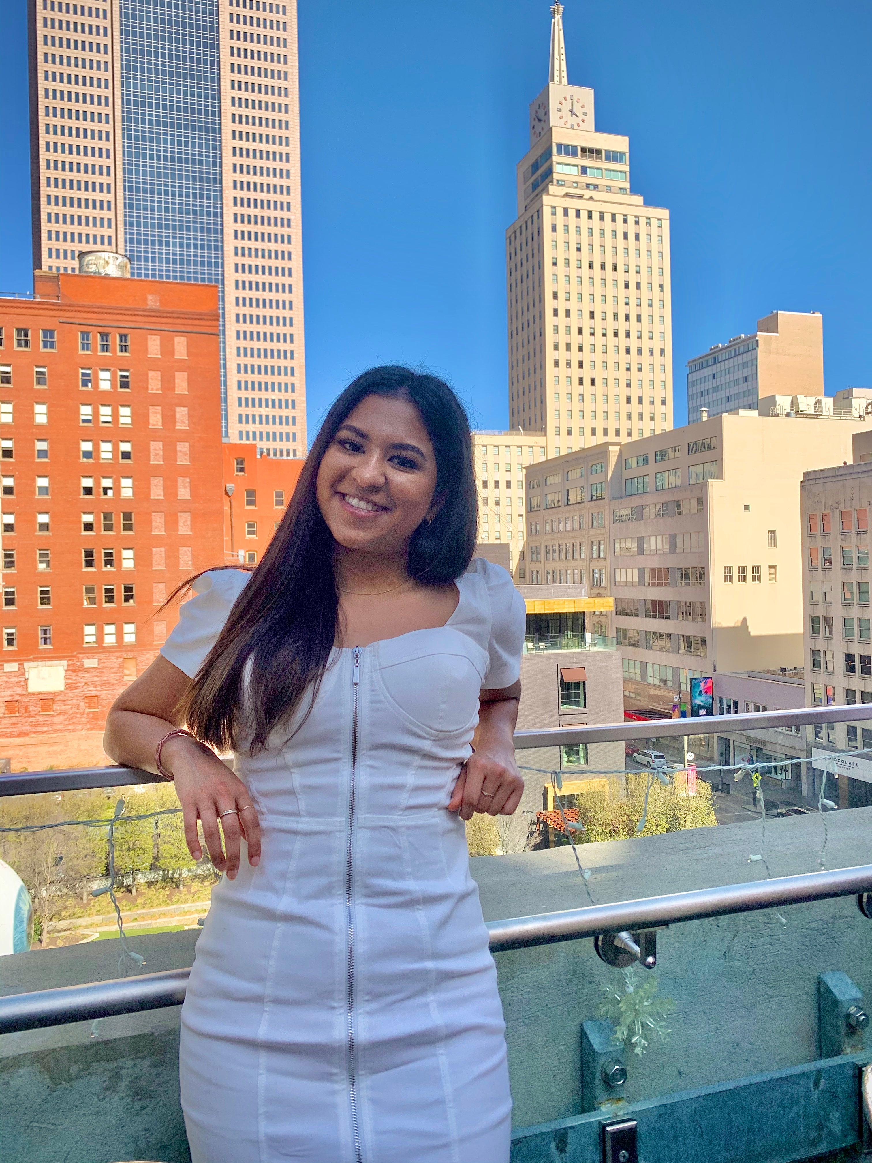 A young woman in a white dress stands on a balcony smiling with a city 's buildings behind her