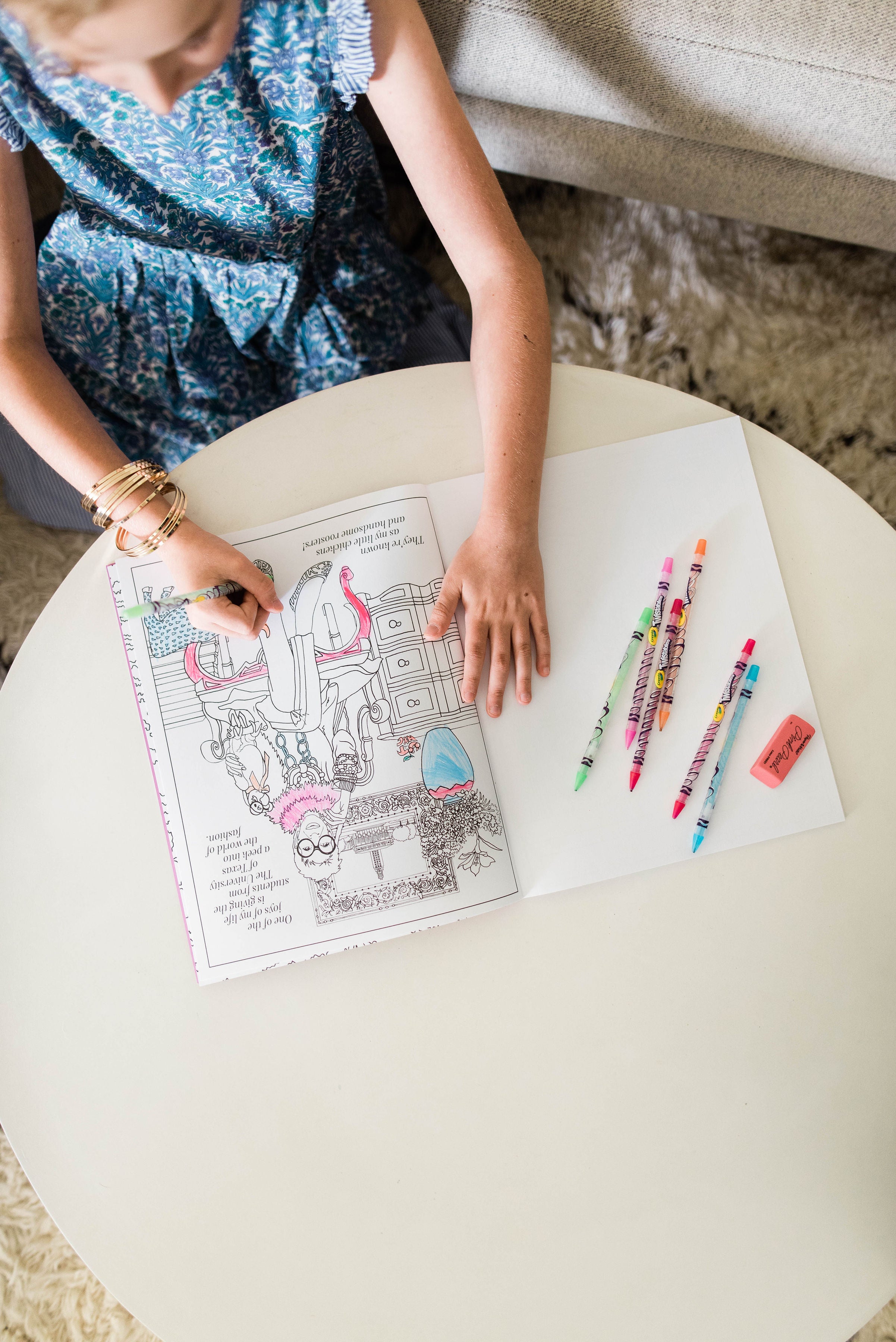 A woman in a blue shirt sits at a round white table using colored pencils to color a page from the coloring book featuring Iris Apfel