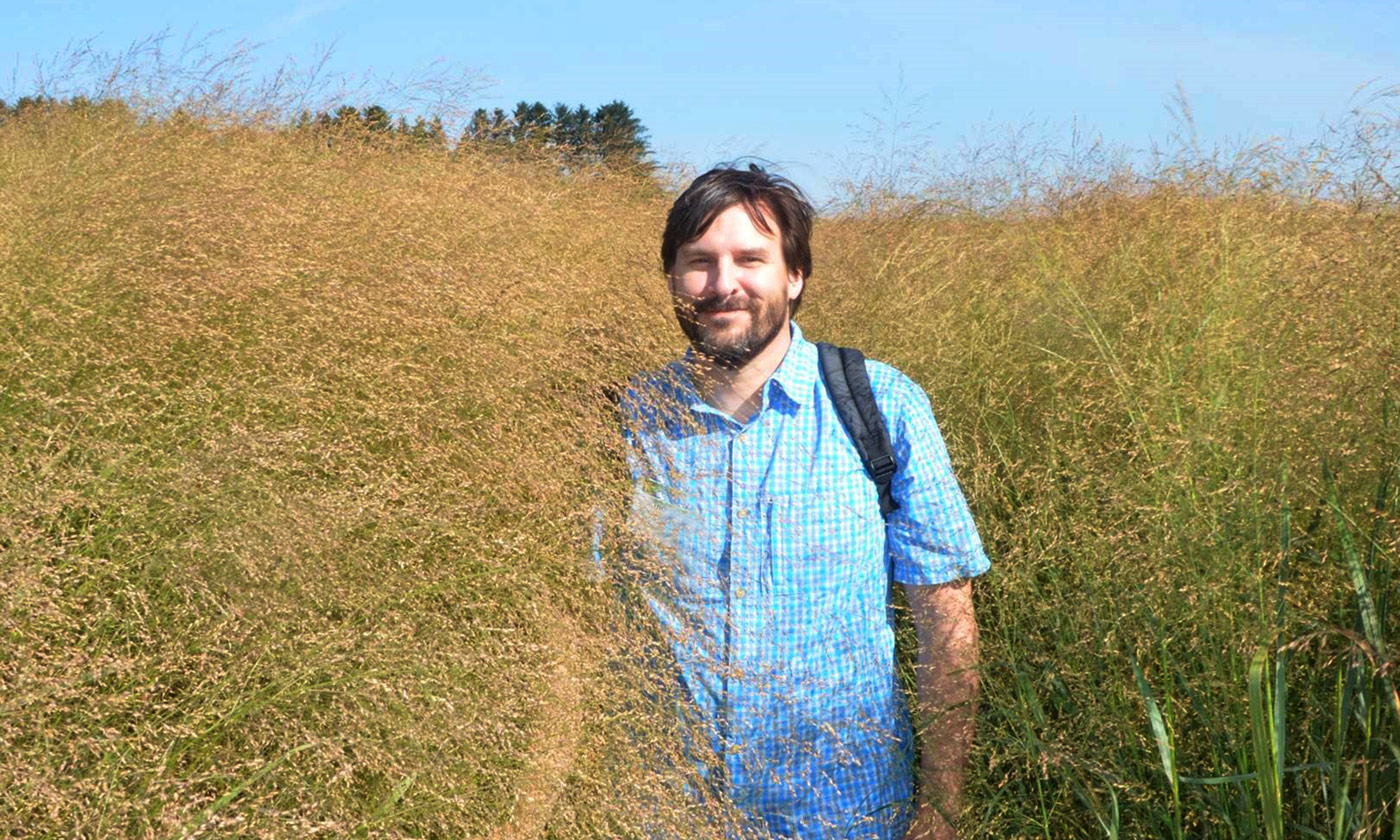 A bearded man in a blue shirt stands in a field of tall grass