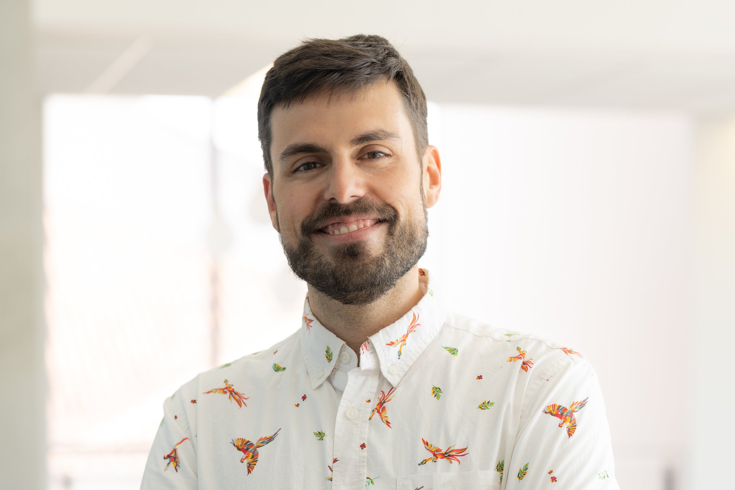 A bearded man in a patterned collared shirt stands in a building in front of a window