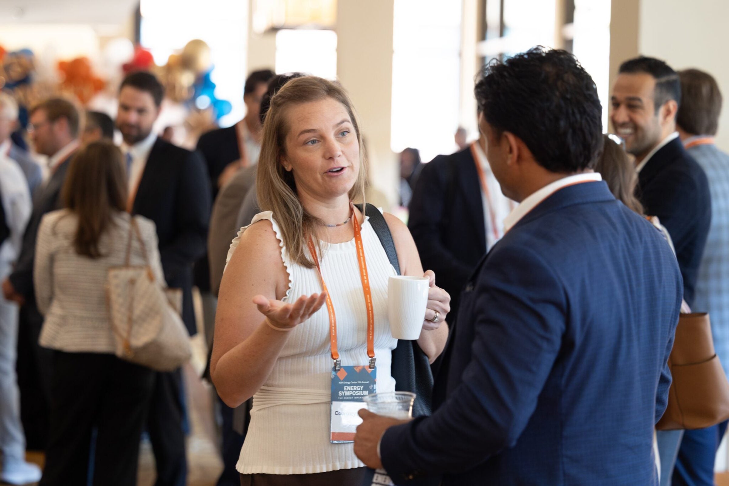 Two people in professional attire talk in a room full of other professionals.