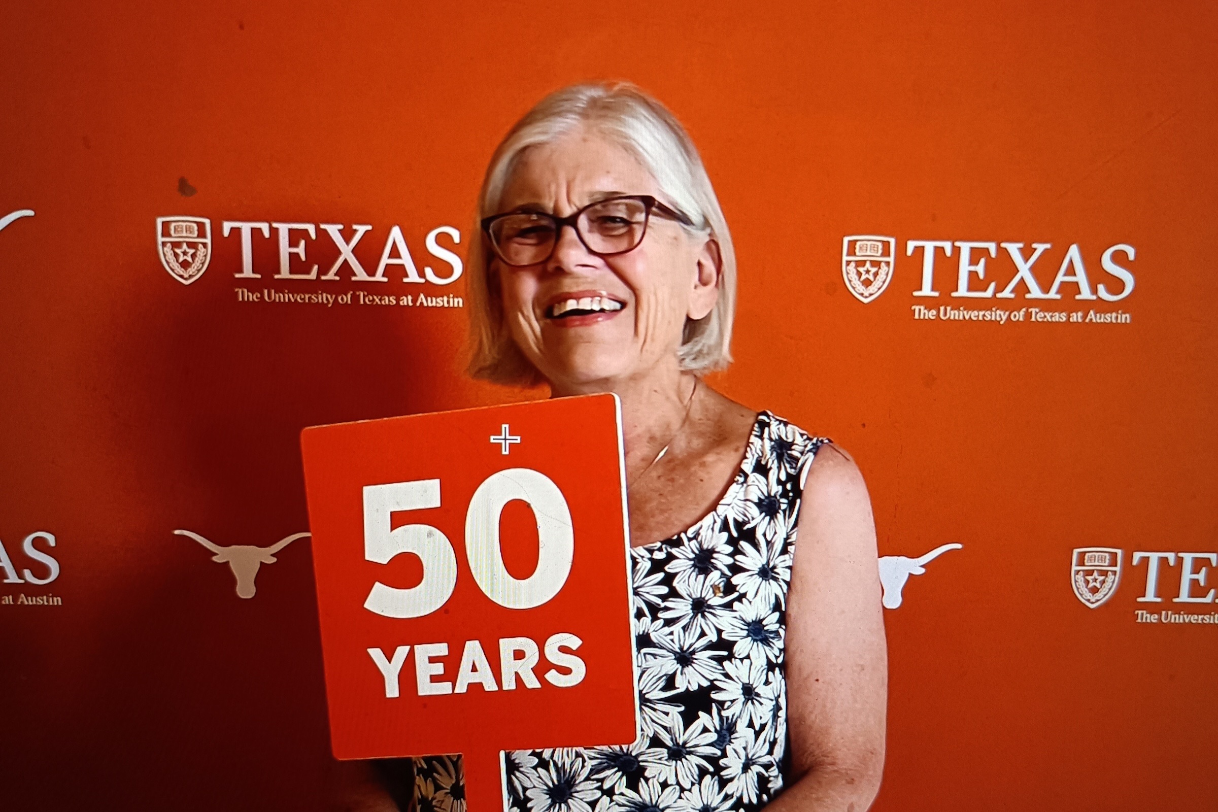 A woman in glasses stands in front of a University of Texas step and repeat sign holding a sign that reads 50 years