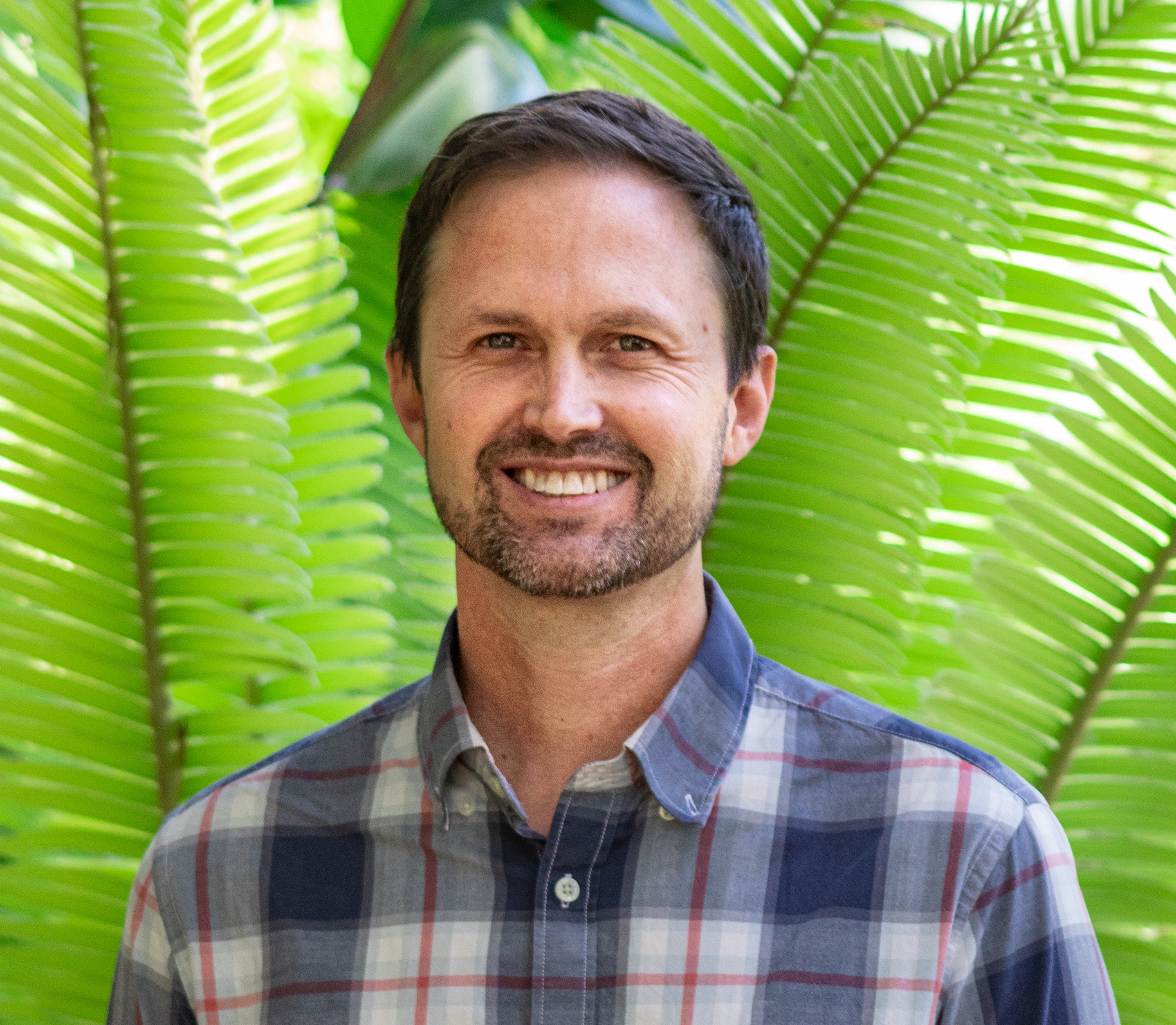 A bearded man in a collared shirt sands in front of a plant with fronds