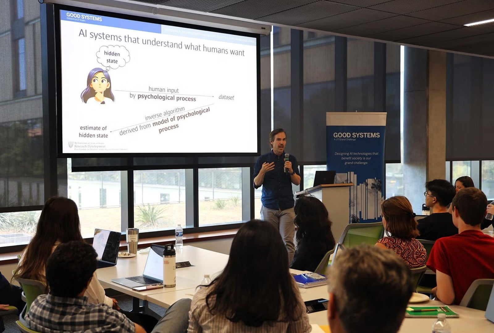 A man holds a microphone and speaks to a group, in front of a banner that reads "Good Systems: A UT Grand Challenge Designing AI technologies that benefit society is our grand challenge" and a slide titled "AI systems that understand what humans want" as a cartoon girl's thought bubble reads "hidden state" and arrows pointing to the words dataset and estimate of hidden state are labeled "human input by psychological process" and "inverse algorithm derived from model of psychological process"