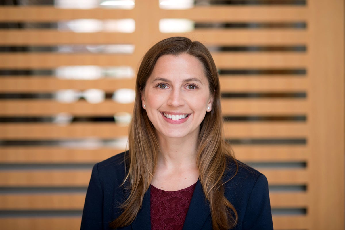 A woman with long hair smiles, standing before a slatted wooden structure indoors.