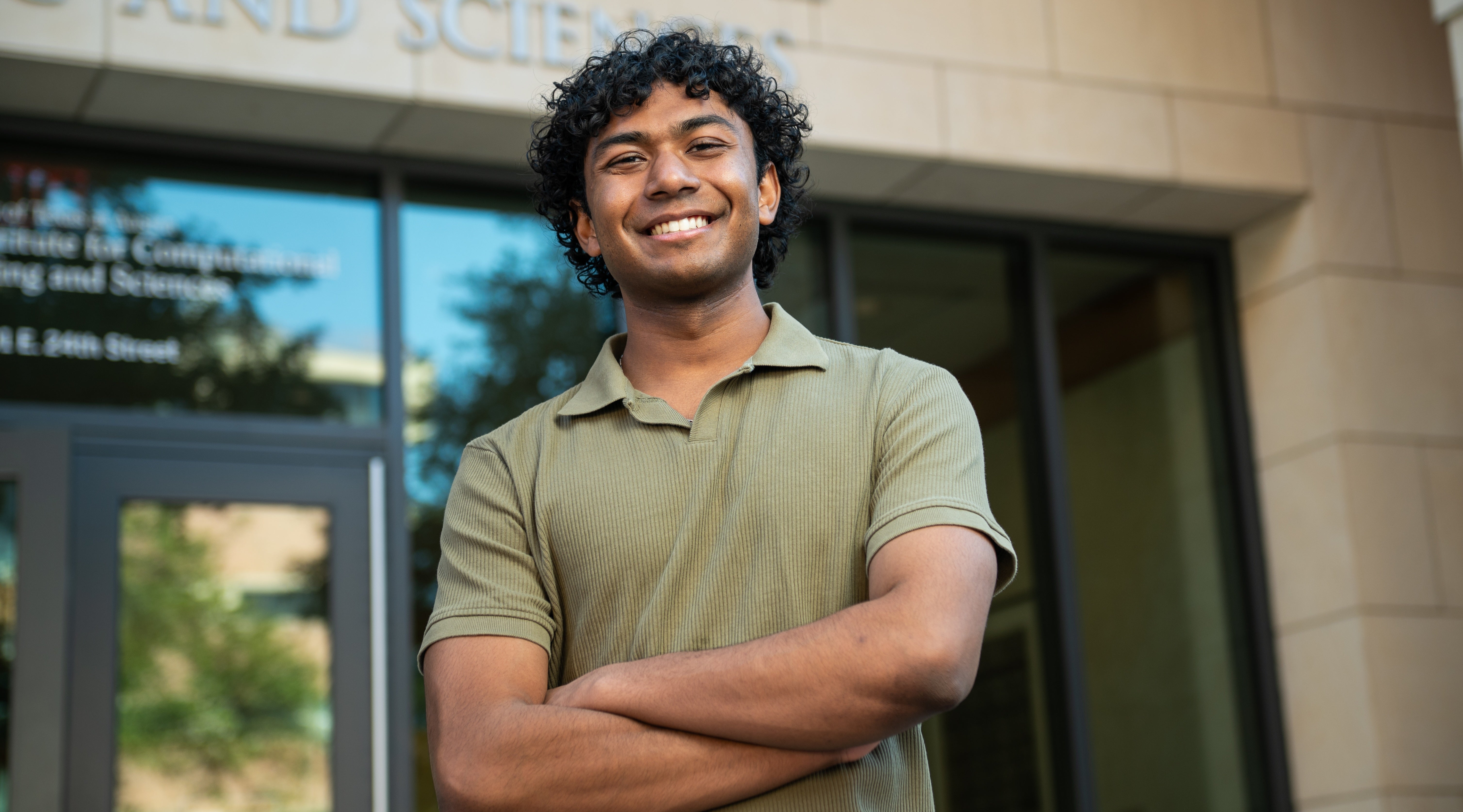 A young man in a polo-style shirt crosses his arms and smiles standing before a limestone building labeled computational engineering and sciences. 