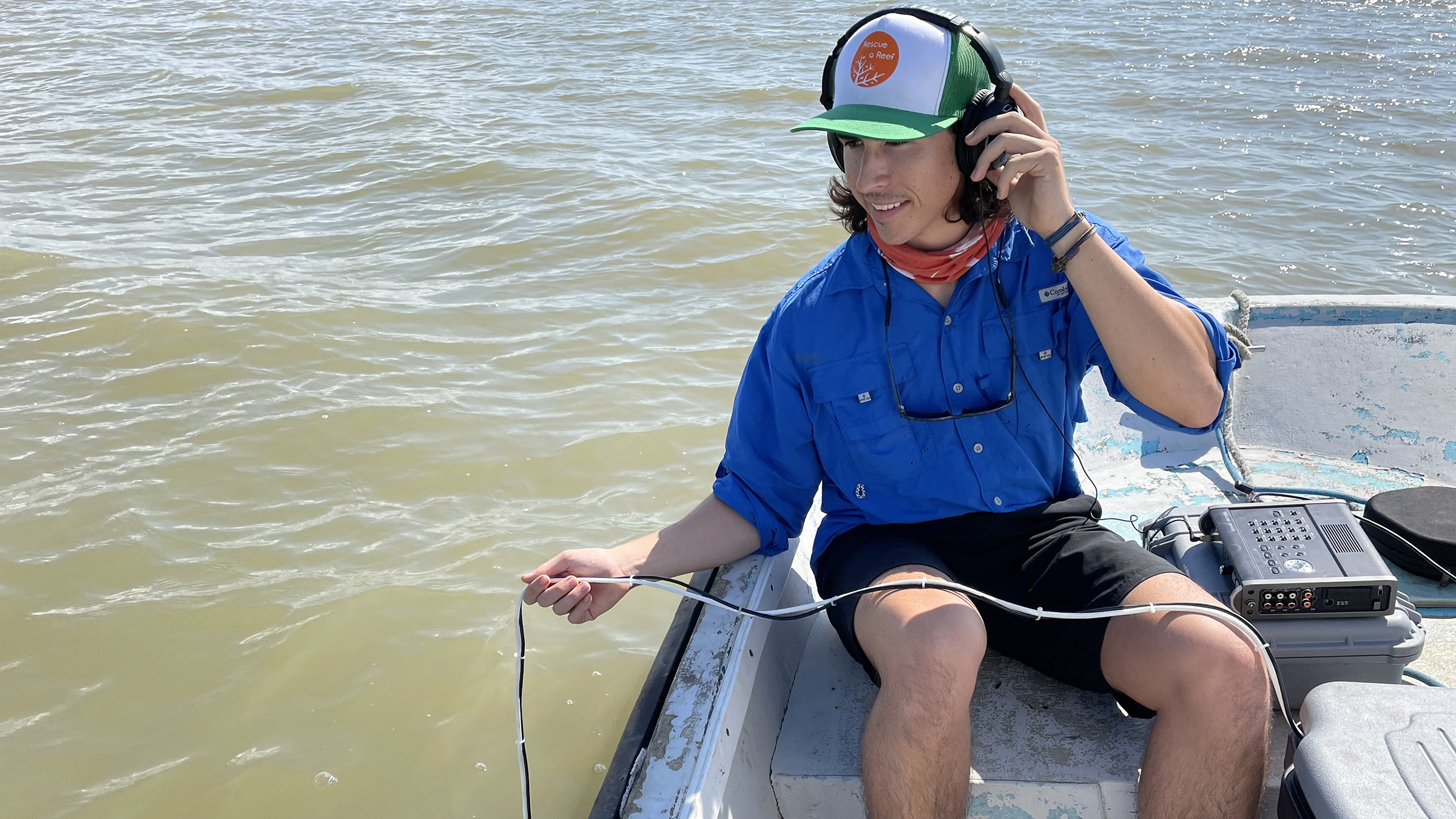 A scientist in a boat wearing headphones and dangles a microphone in the water