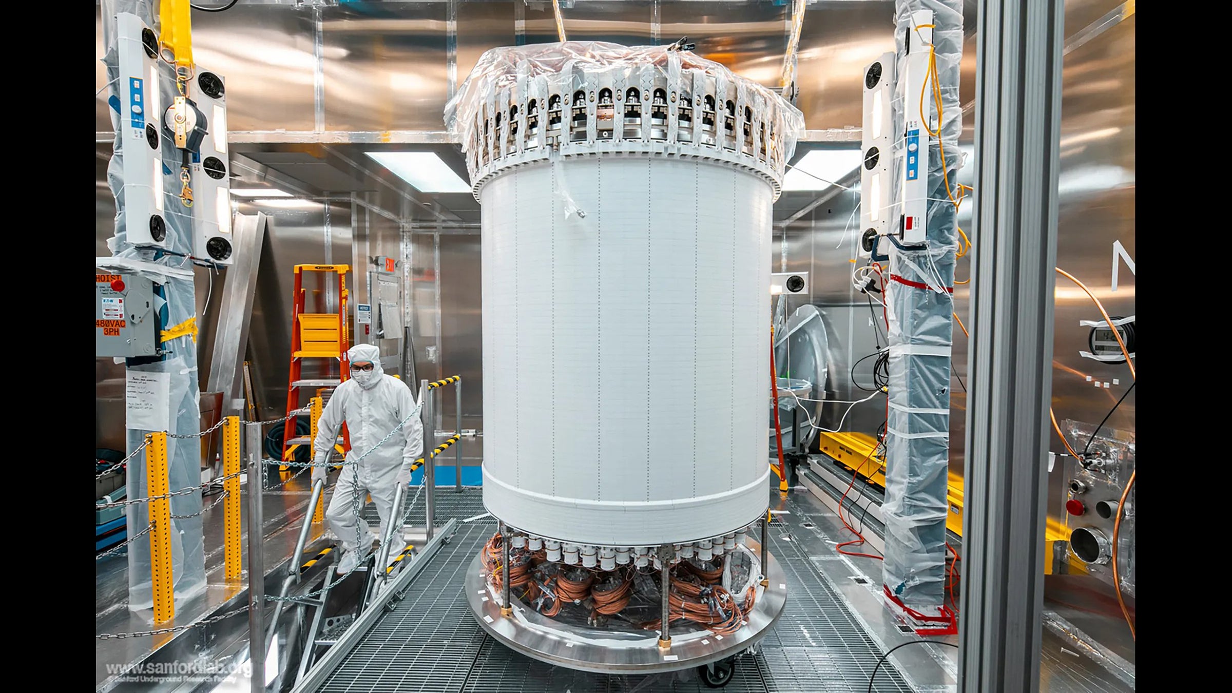 A large round white cylinder sits in the center of a laboratory and a worker wearing a white clean room suit walks down stairs nearby.