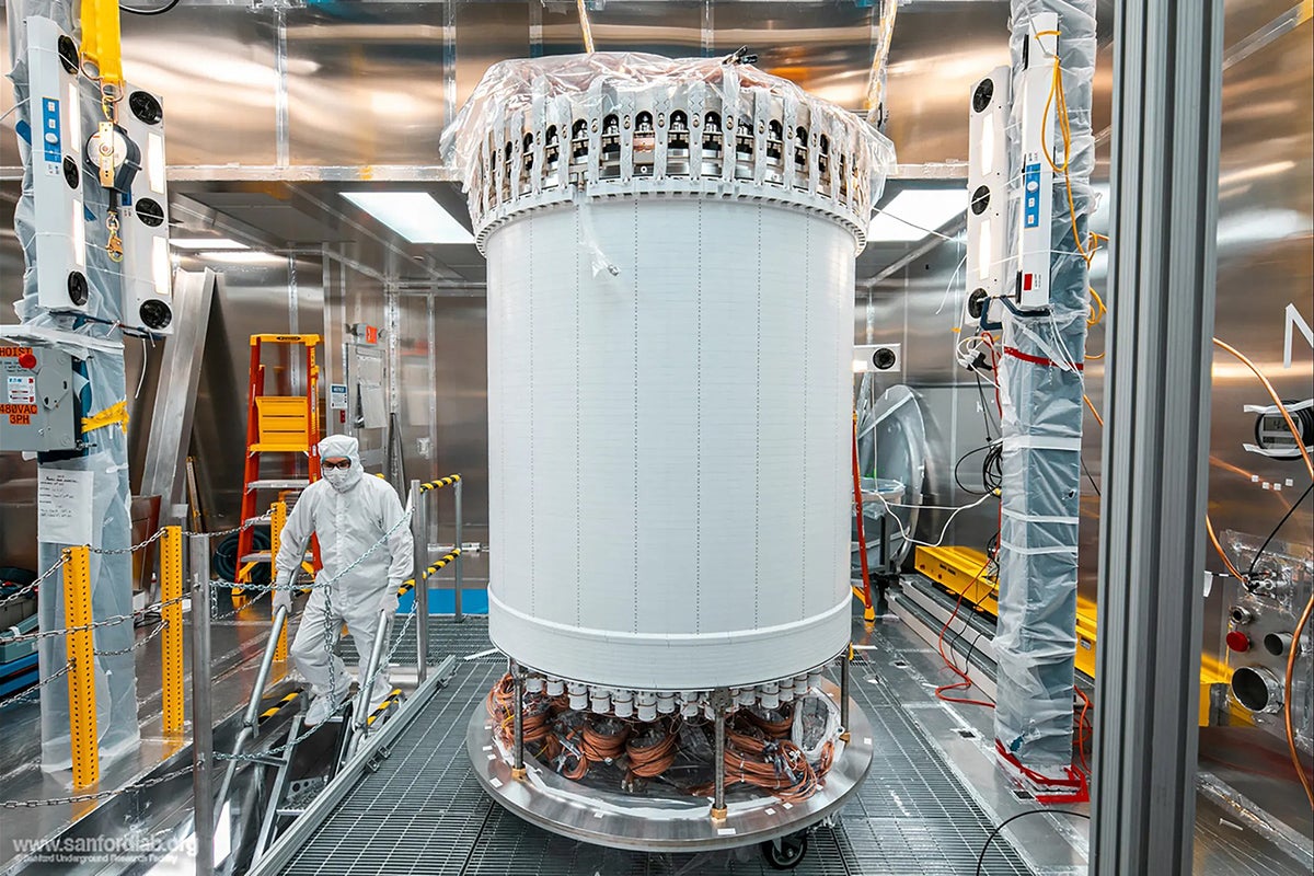 A large round white cylinder sits in the center of a laboratory and a worker wearing a white clean room suit walks down stairs nearby.
