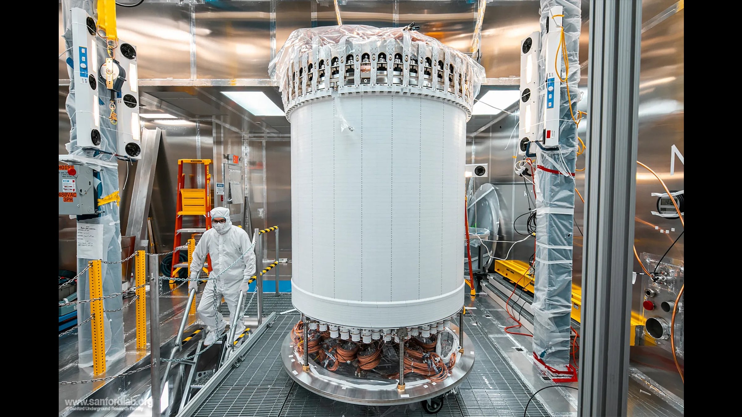 A large white cylinder in the center of a laboratory. A person wearing a full-body white clean room outfit descends nearby stairs.