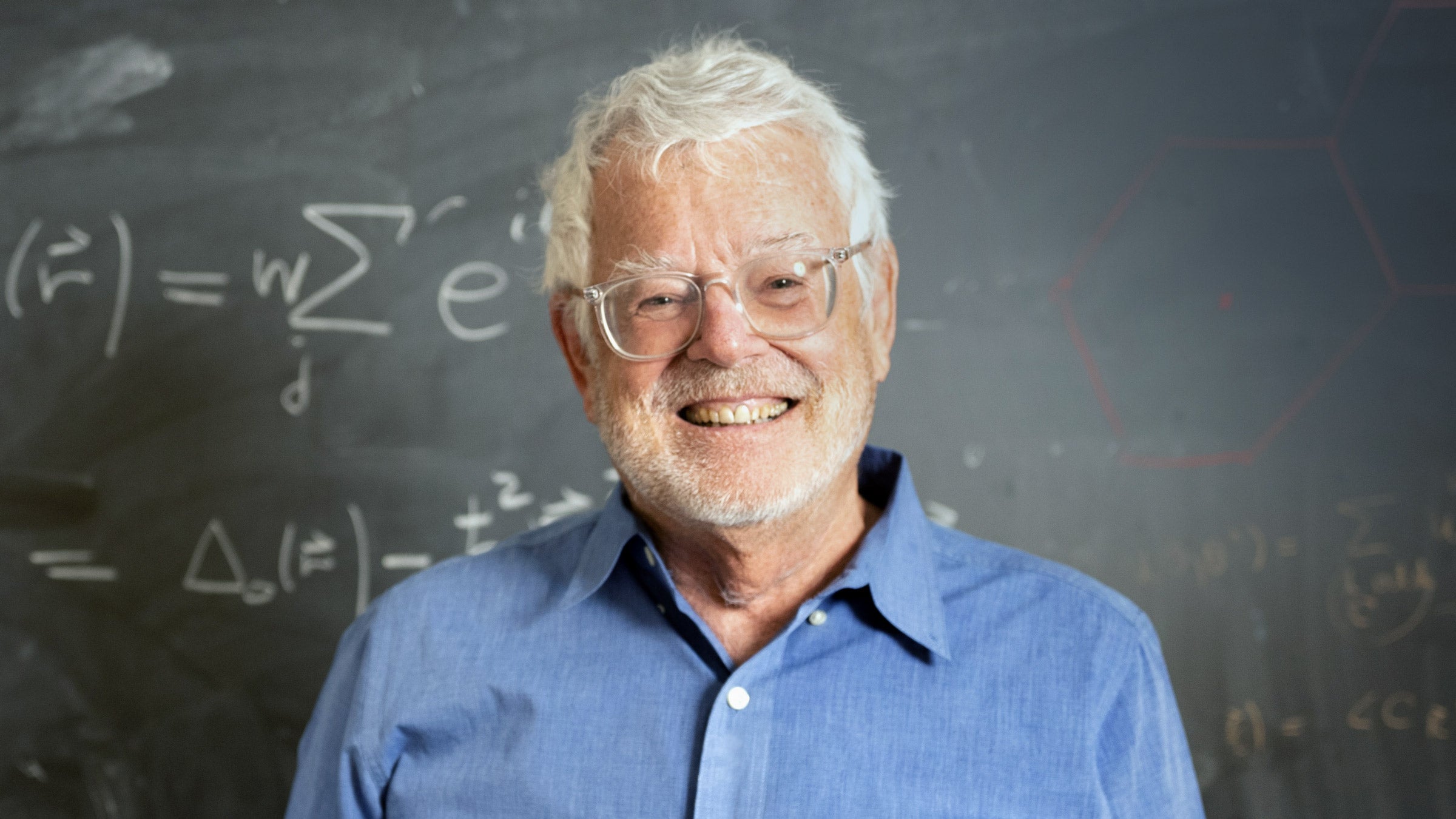 A man in a collared shirt and glasses smiles, standing before a chalkboard with equations.