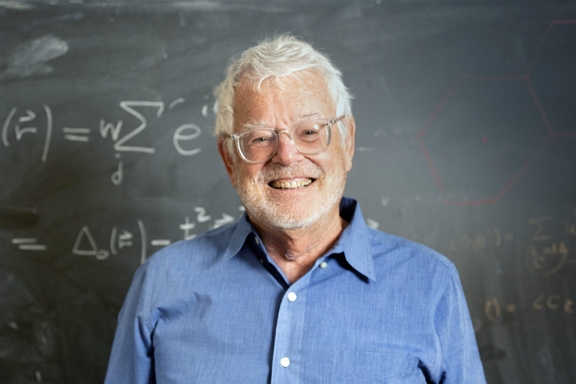 A man in a collared shirt and glasses smiles, standing before a chalkboard with equations.