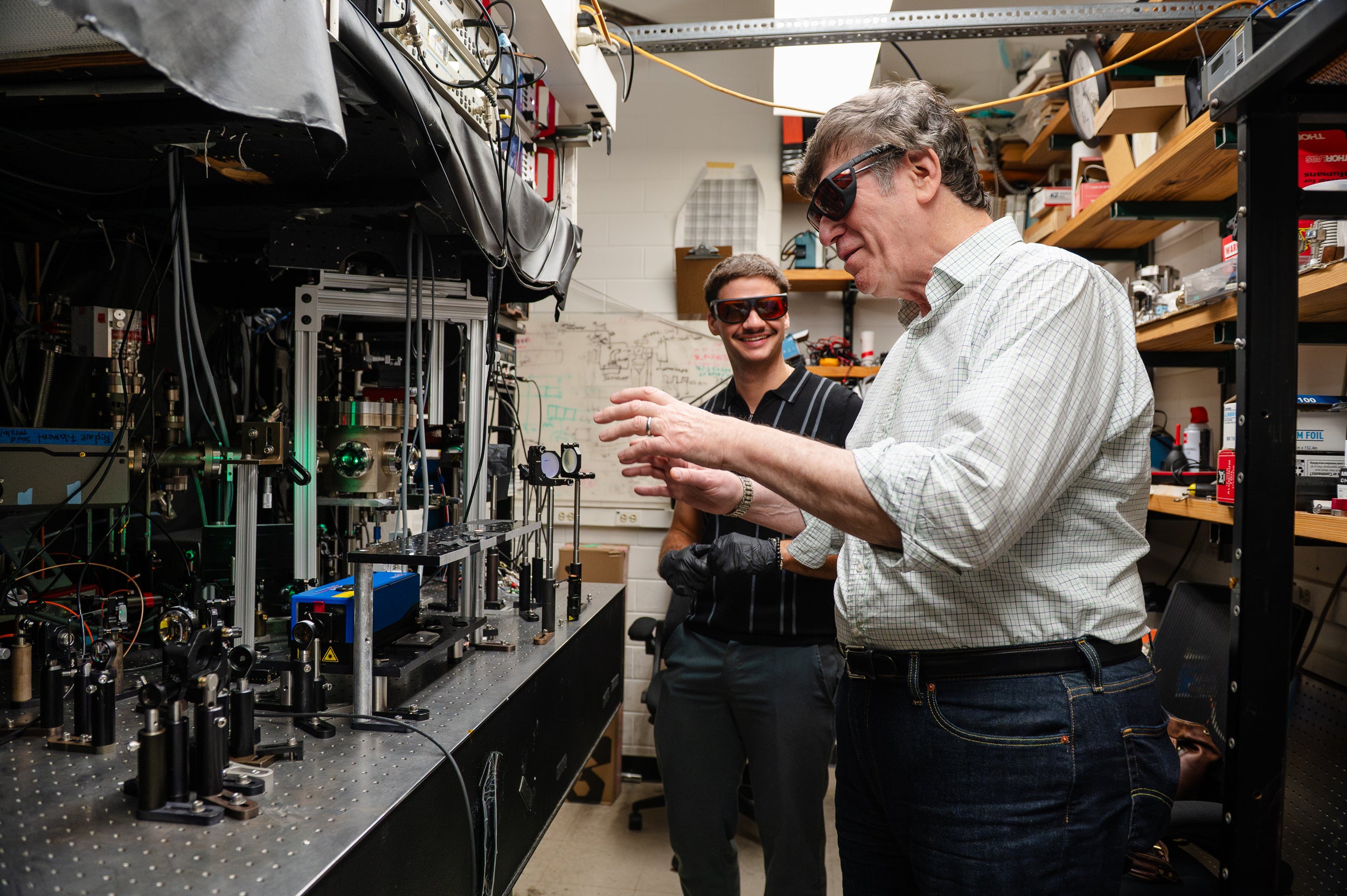 Two scientists wearing eye protection stand in a lab before some laser equipment, as one gestures at the device.