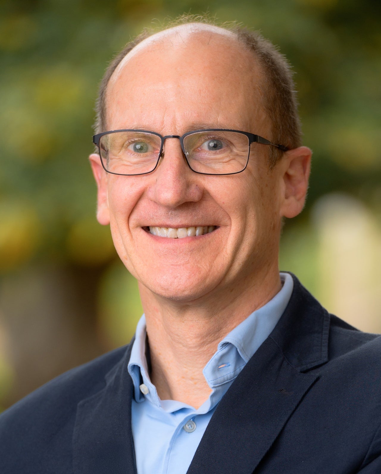 A man in glasses and a suit jacket smiles in front of a verdant background.