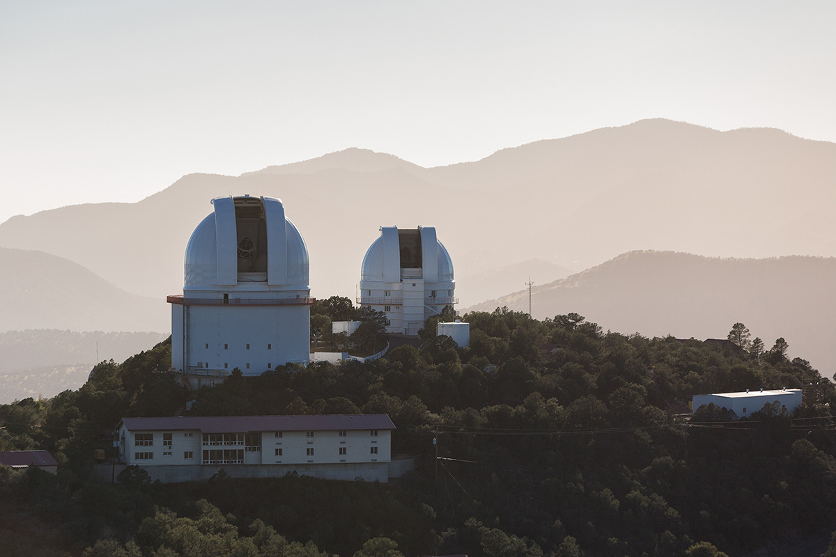 Two telescope domes sit atop a mountain with more peaks in the distance