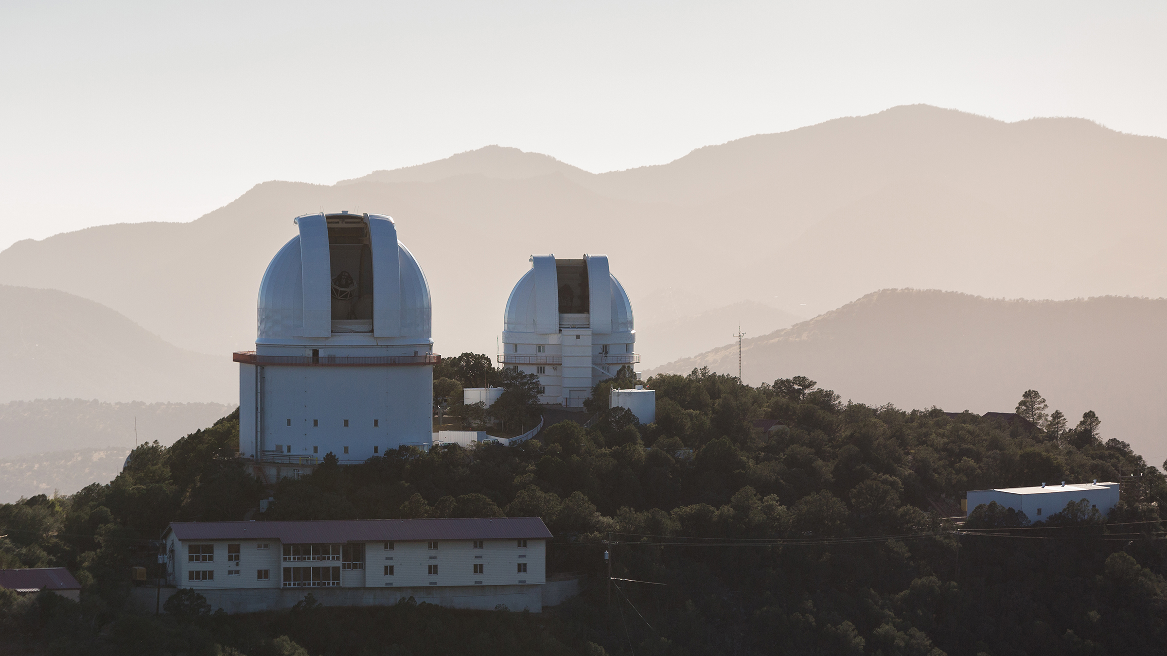 Two telescope domes sit atop a mountain with more peaks in the distance