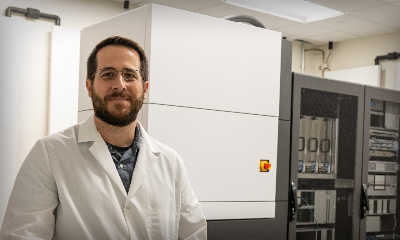 A bearded man in a white lab coat stands in front of a large white cabinet