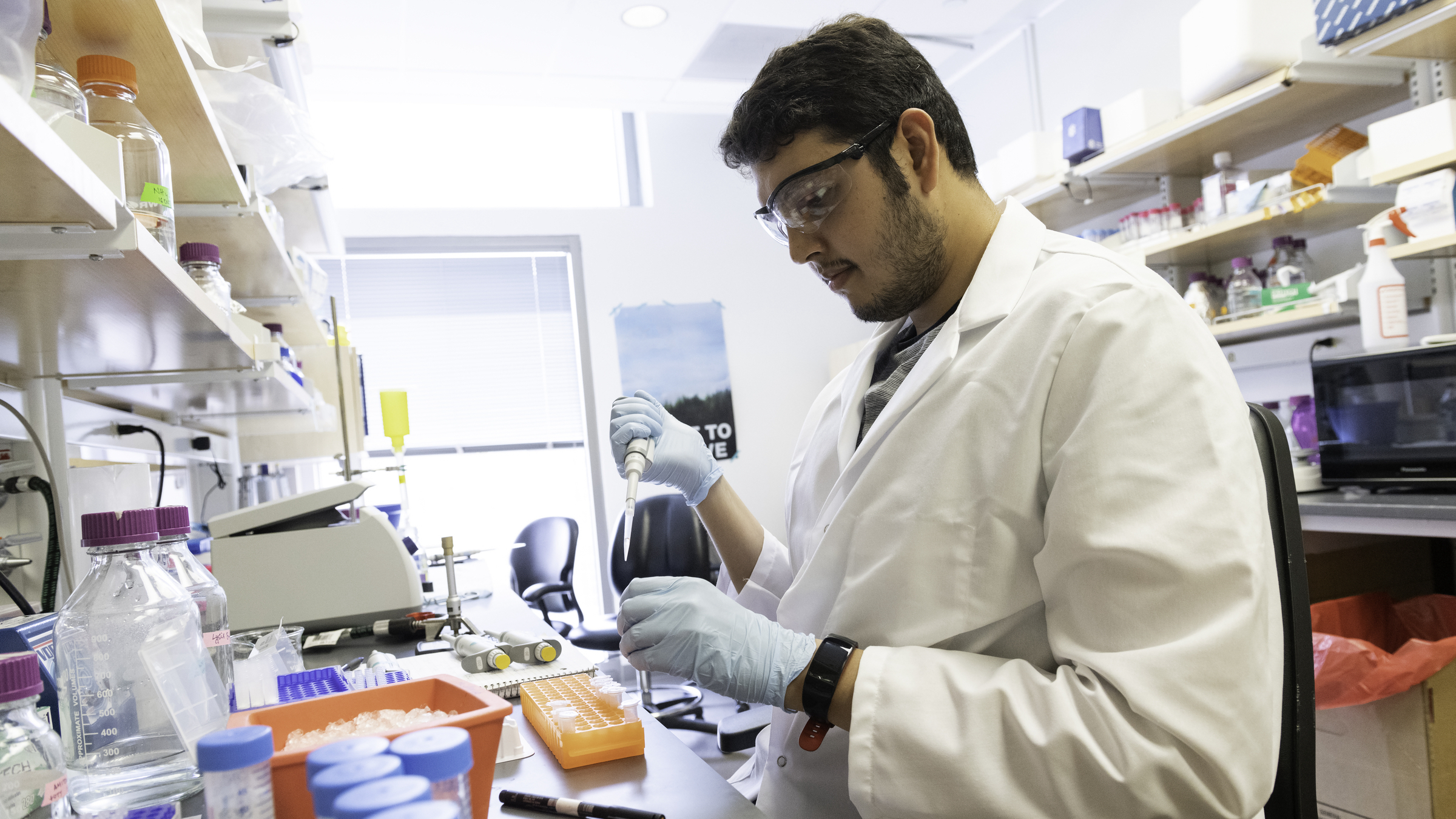 A graduate student in a white coat pipettes in a lab wearing gloves