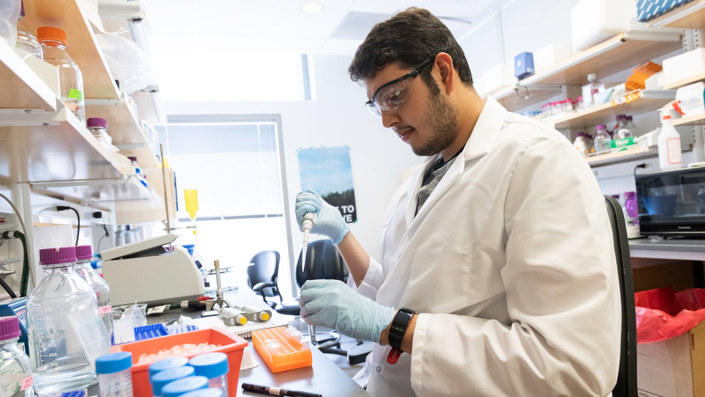 A graduate student in a white coat pipettes in a lab wearing gloves