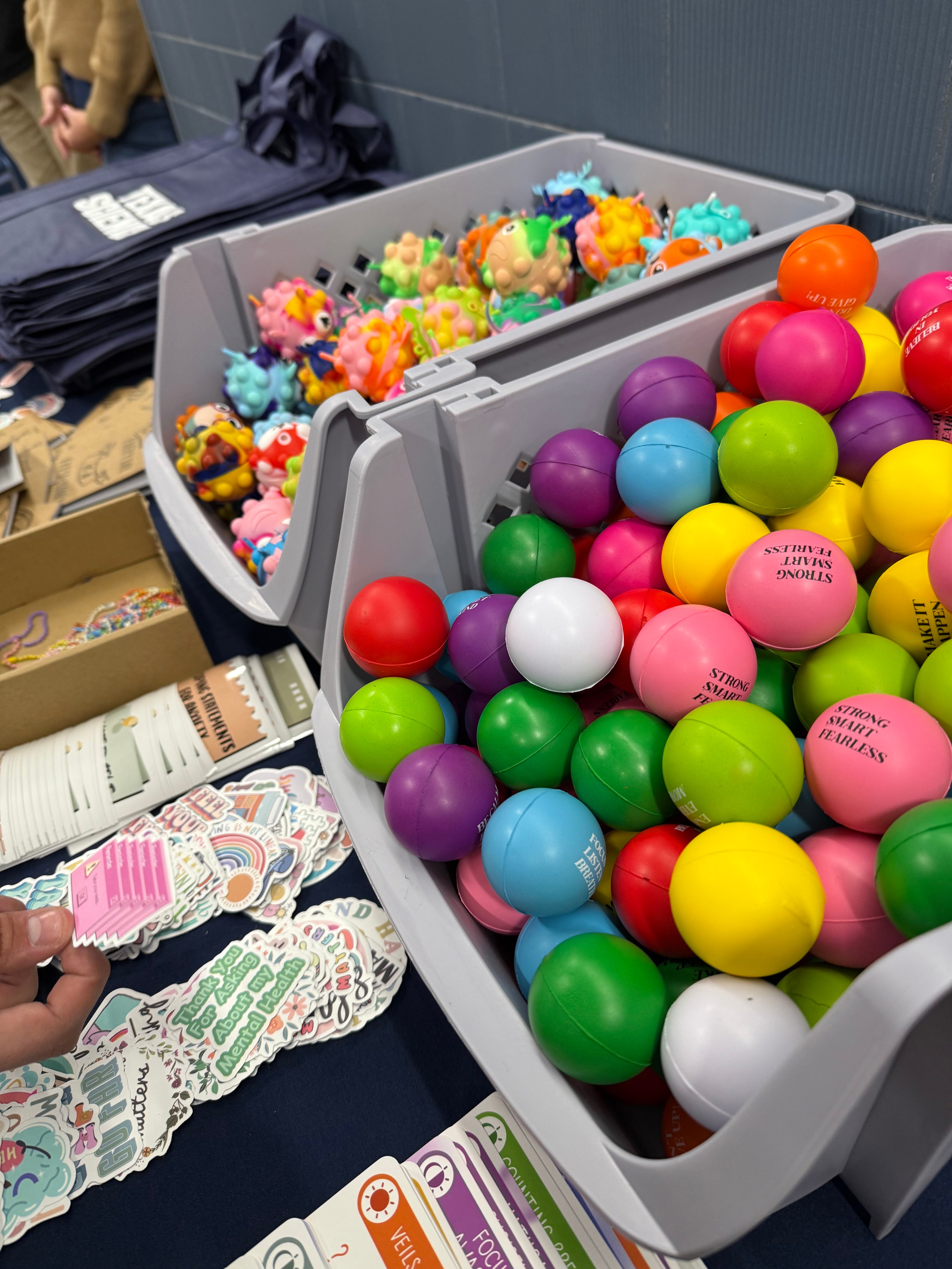 A hand holds a sticker up from a group of giveaway stress balls and stickers, some of which read "thank you for asking about my mental health"