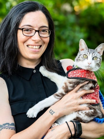 A smiling woman with black-rimmed glasses and dark hair holds a cat with calico and grey patches along its head and back and white fur underneath. The cat is wearing a red safety harness.