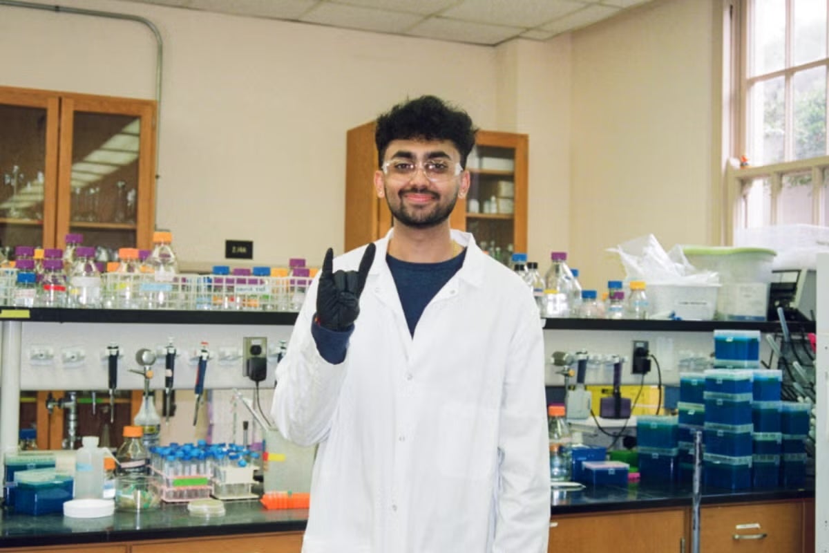 A young man with a beard wears PPE in a wet lab and flashes the hook 'em horns hand sign.