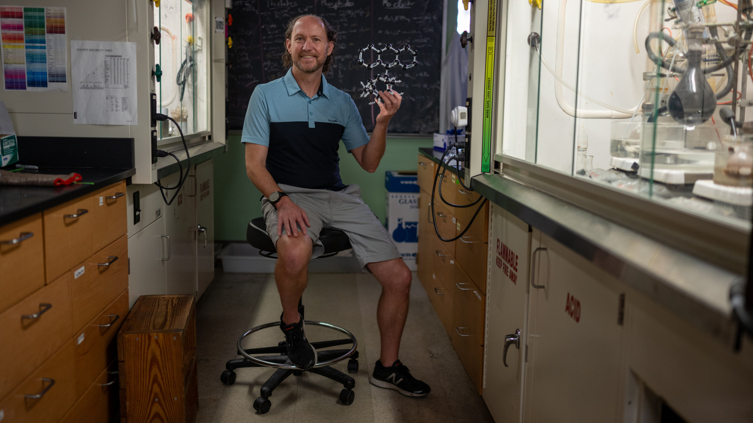 Michael Rose sits between lab benches on a rolling stool holding up a model of a molecule