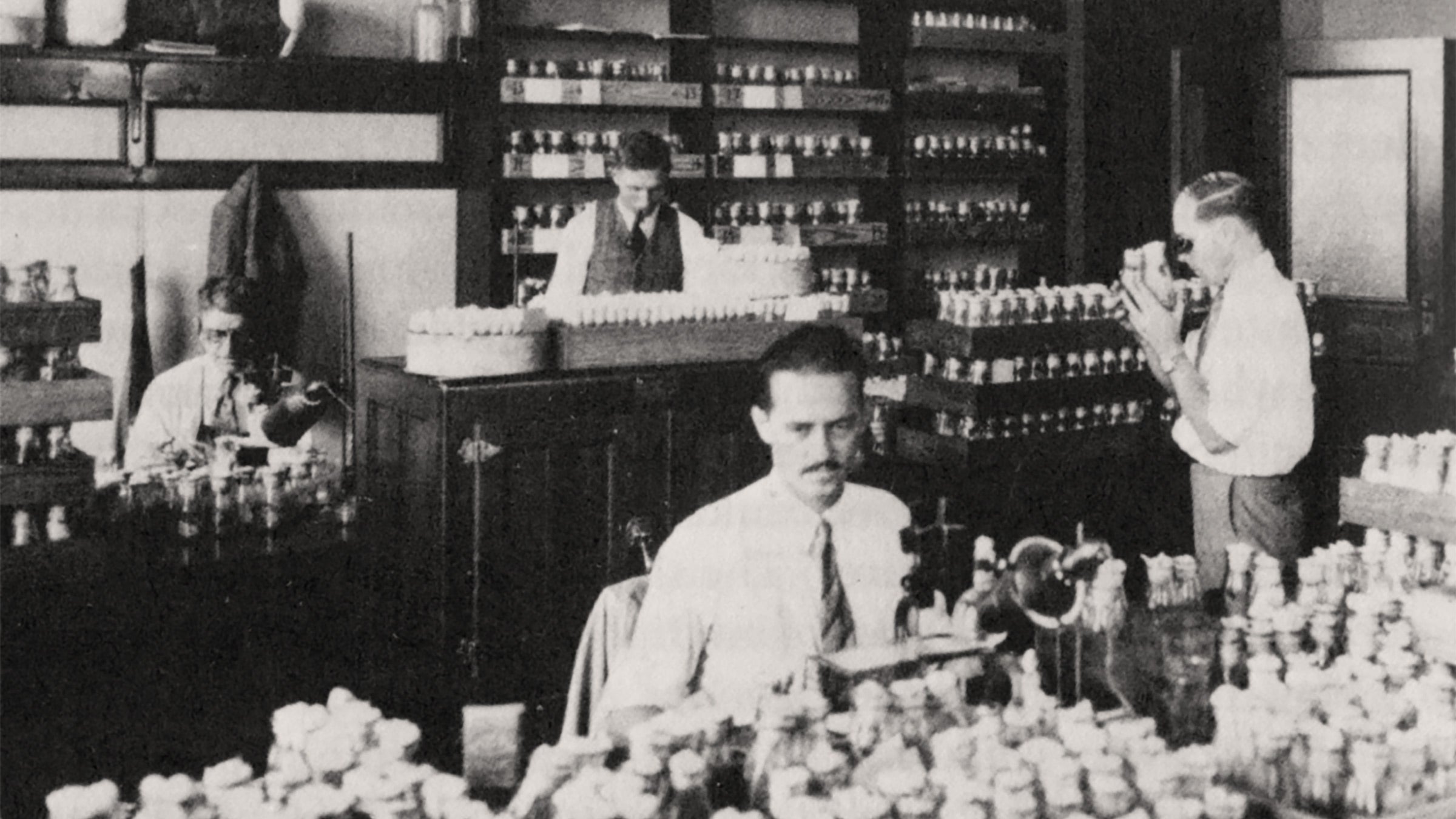Black and white photo of four men working in a lab, wearing white button-up shirts and ties. There are many glass bottles on tables and shelves with white cloth stoppers.