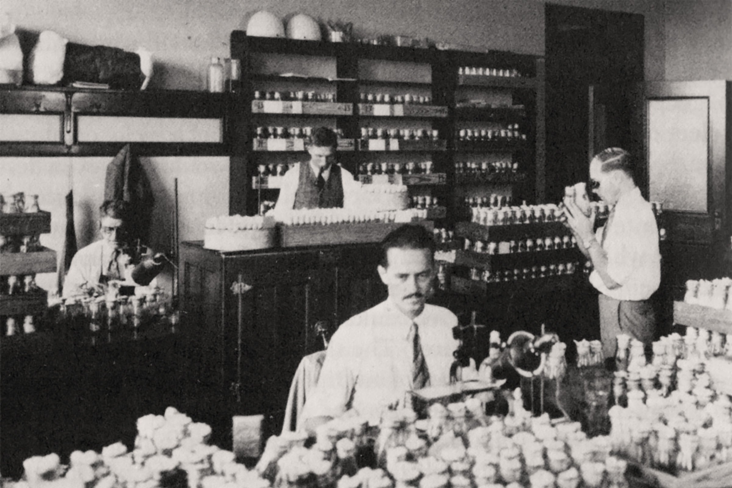 Black and white photo of four men working in a lab, wearing white button-up shirts and ties. There are many glass bottles on tables and shelves with white cloth stoppers.