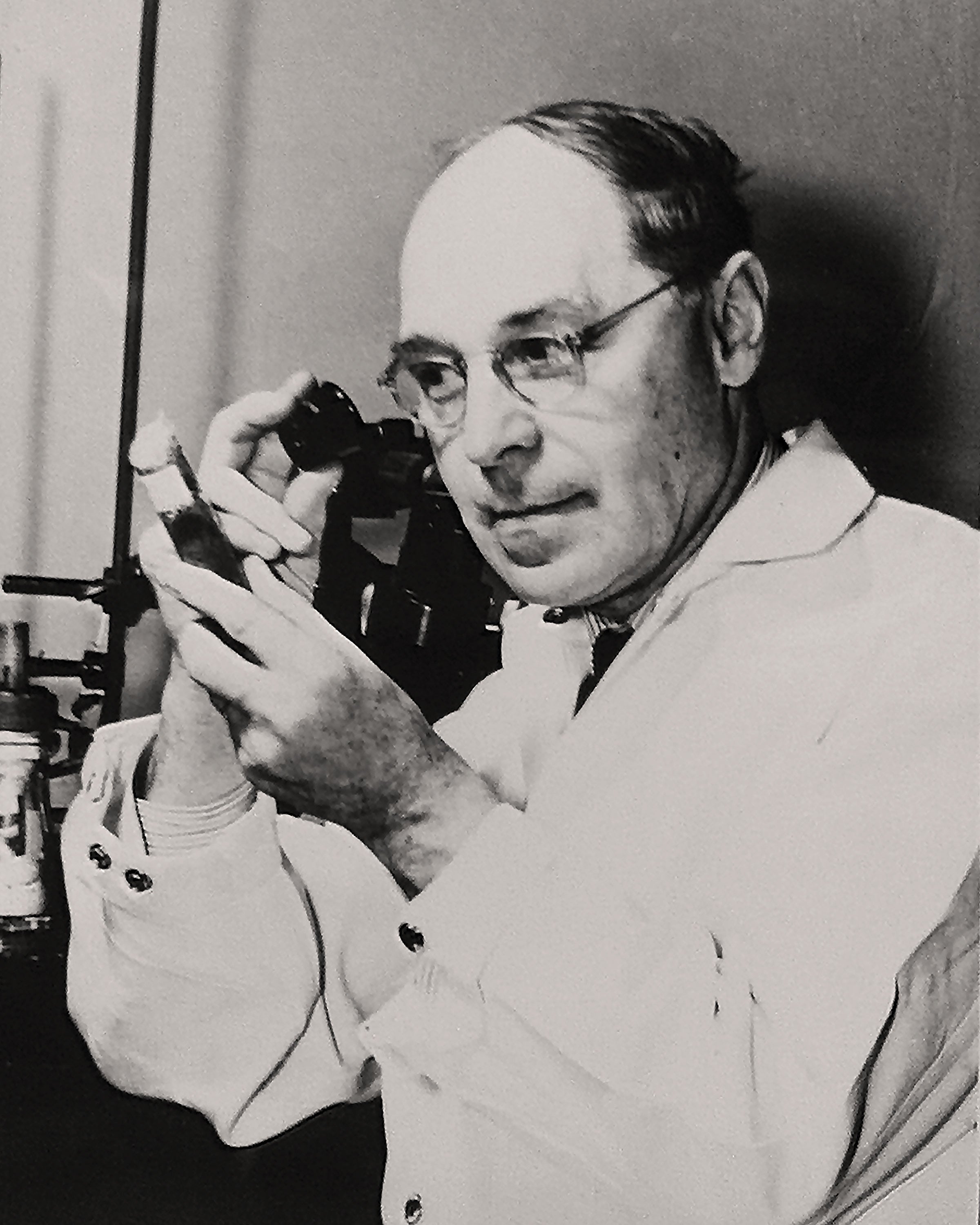 Black and white photo of a middle aged man looking at a glass tube through a jeweler's loupe