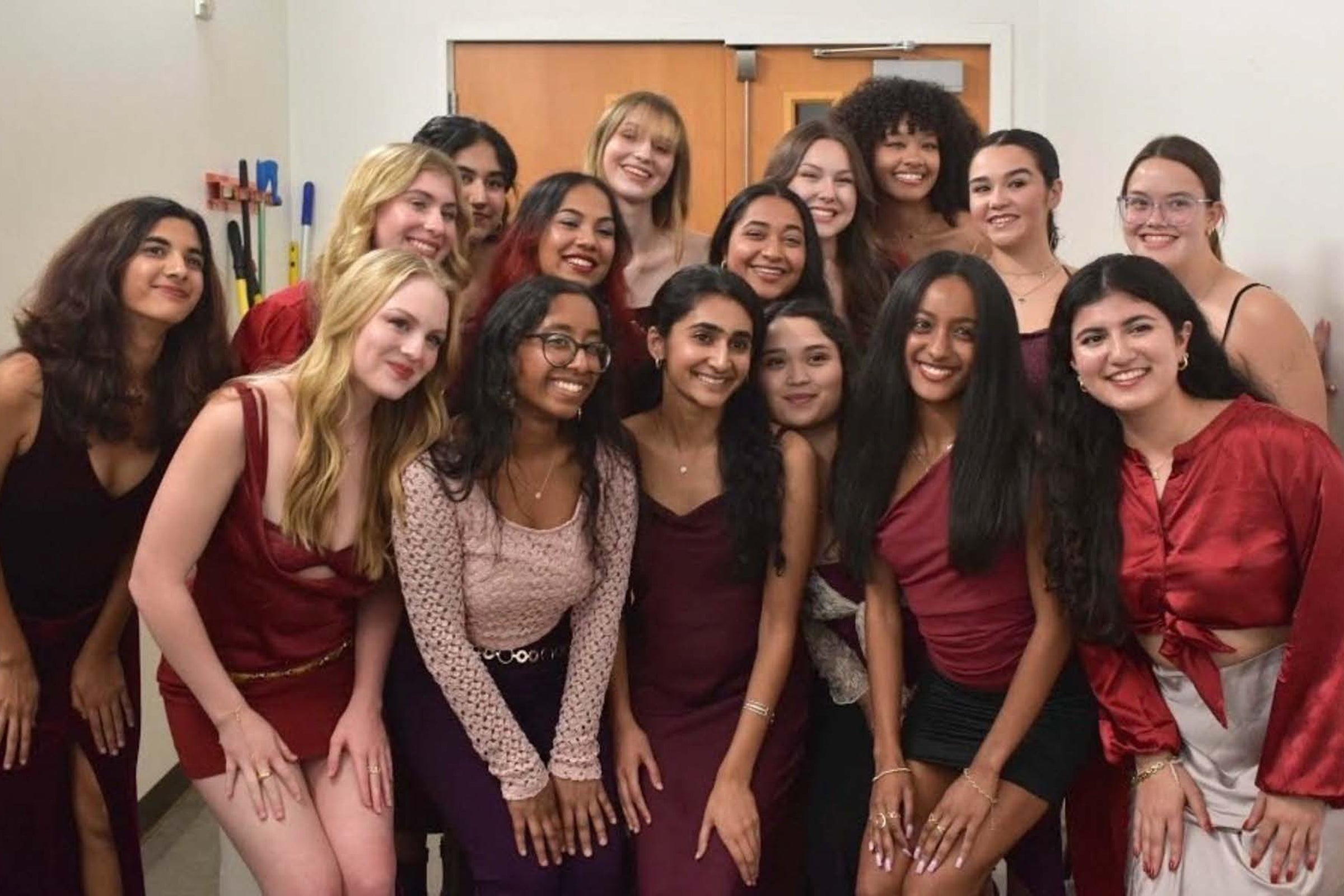 Sixteen smiling women dressed in evening wear huddle together for a group selfie.