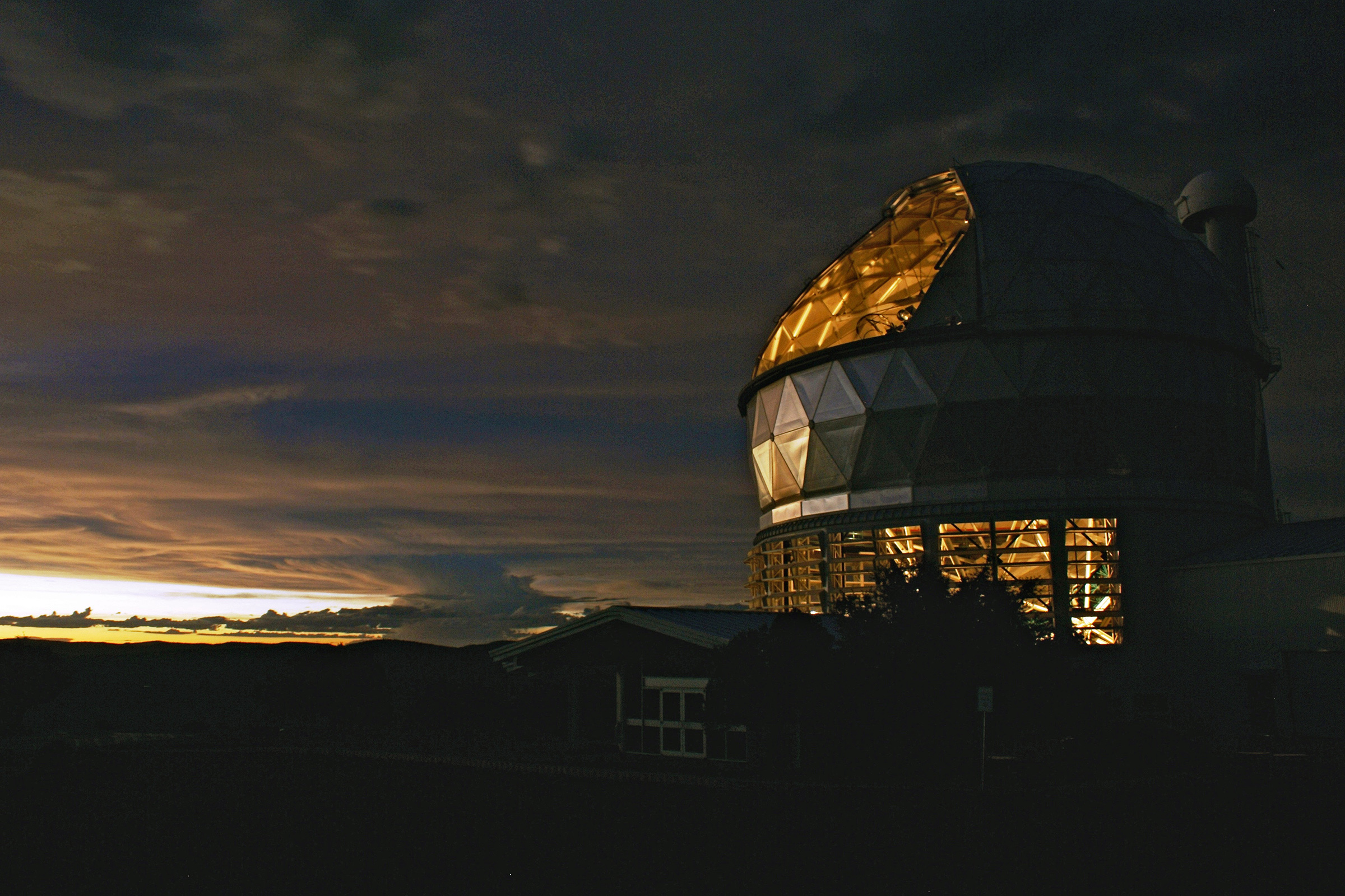 The Hobby-Eberly Telescope is lit from within at dusk as clouds loom in the background