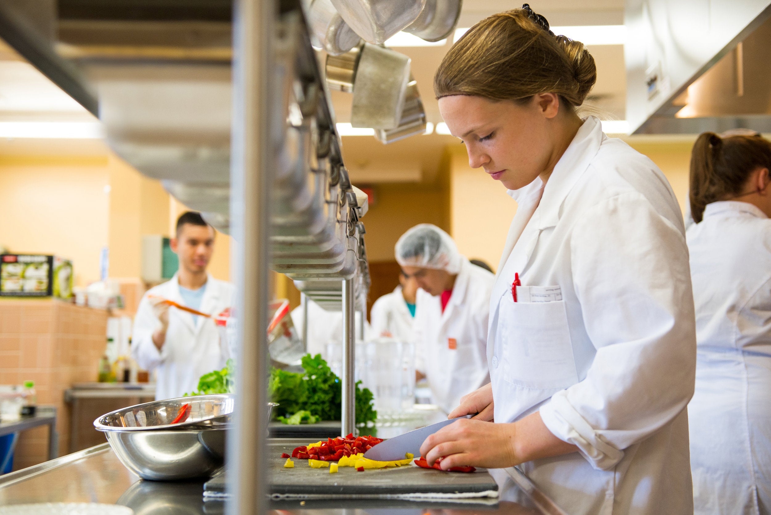 A young woman in a white jacket chops vegetables while others around her do likewise.
