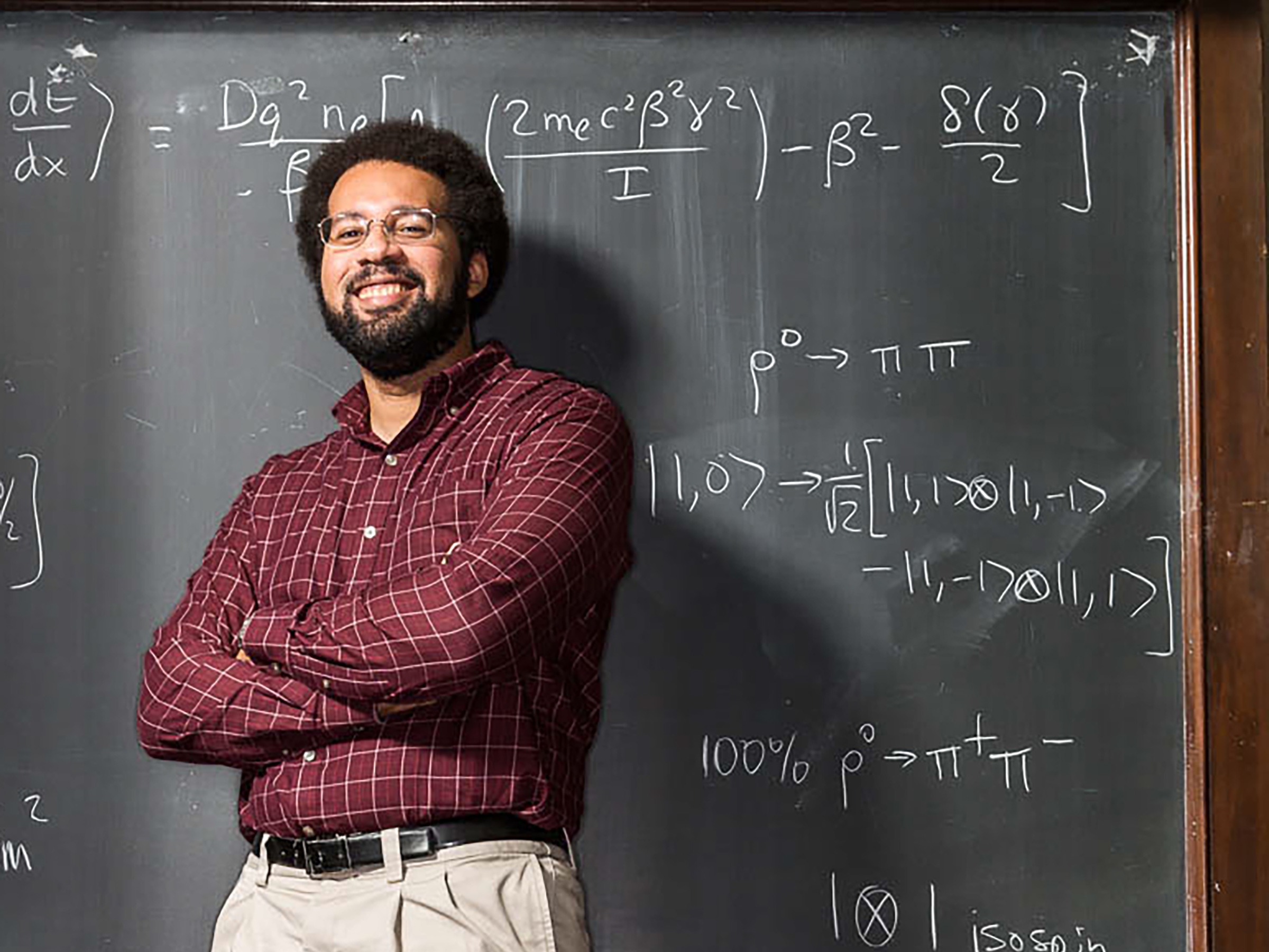 A man stands with arms crossed in front of a chalk board with mathematical equations