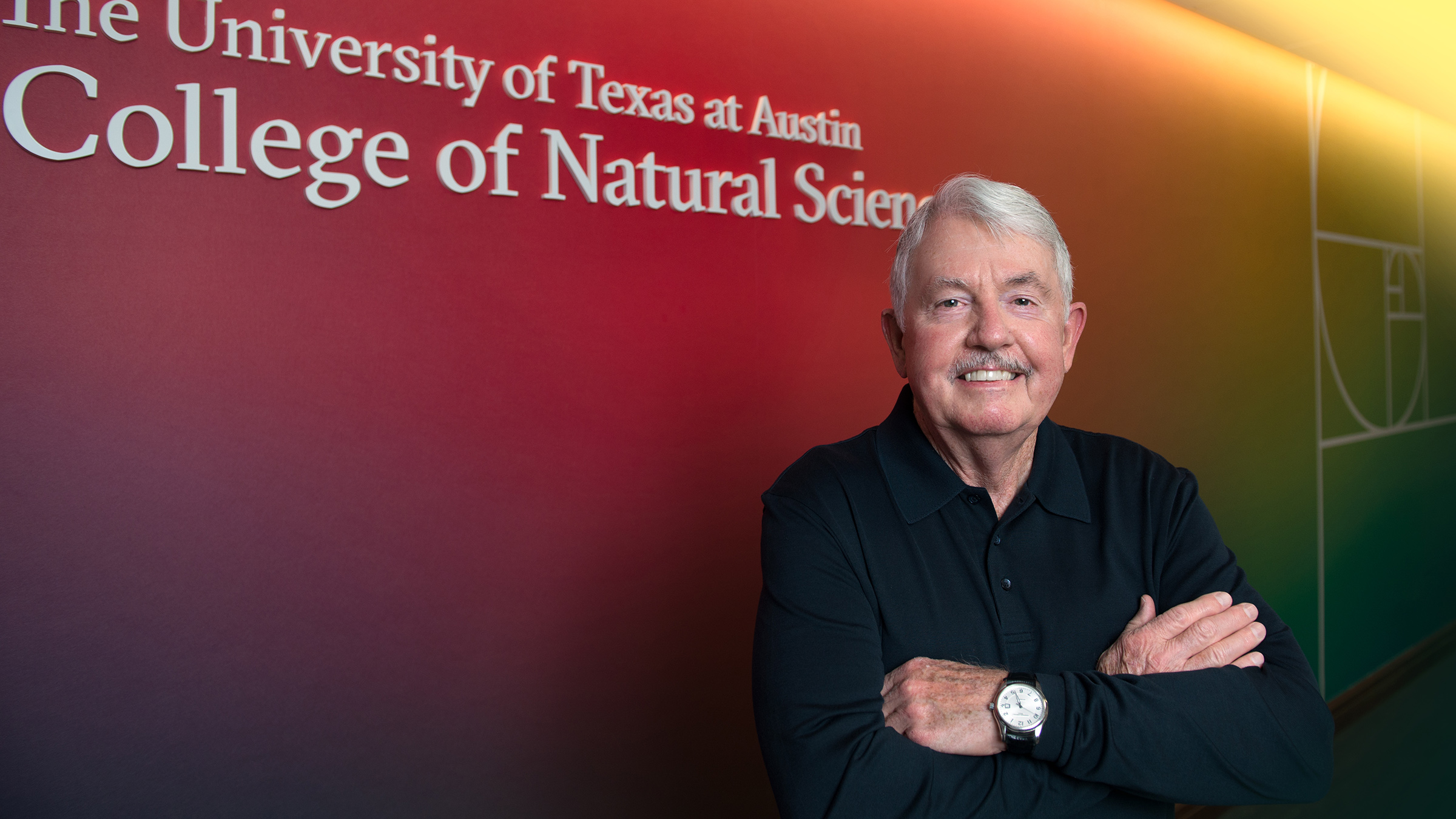 A man stands with arms crossed in front of a wall that says &quot;The University of Texas at Austin College of Natural Sciences&quot;
