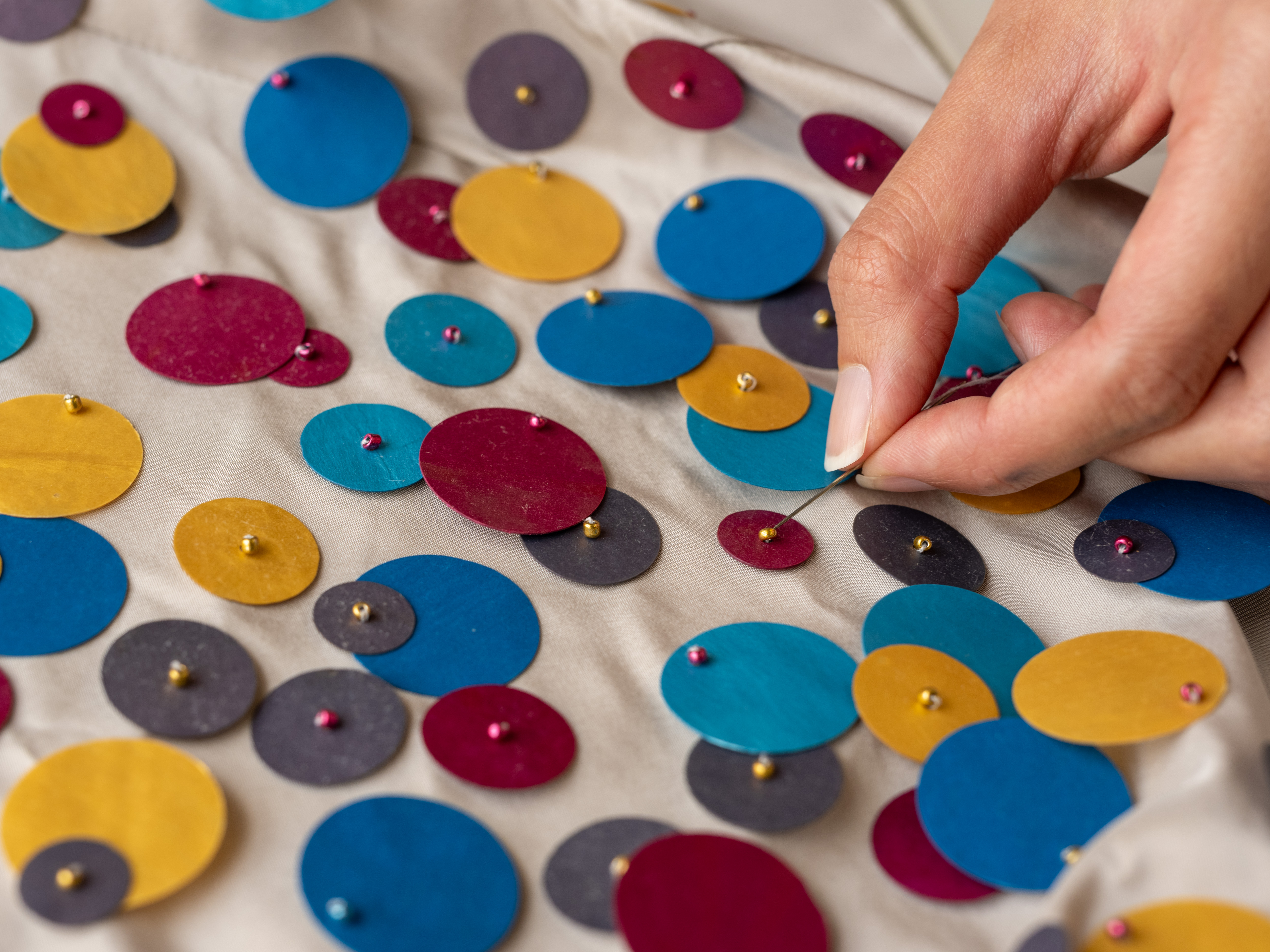 A hand sews a bead onto a cloth with sequins of differing sizes and colors.