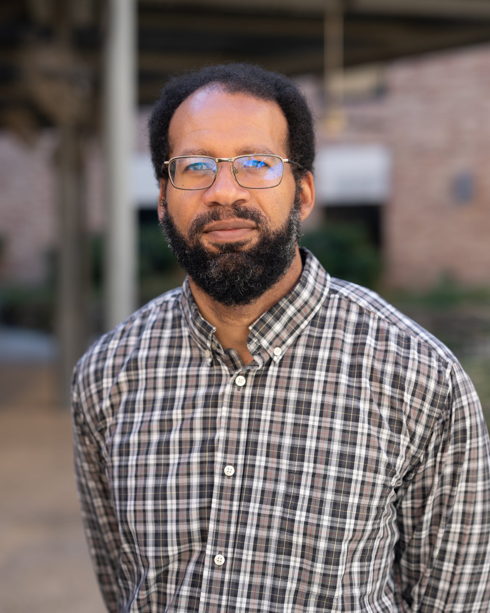 A bearded man in glasses and a button-up plaid shirt stands in a courtyard.