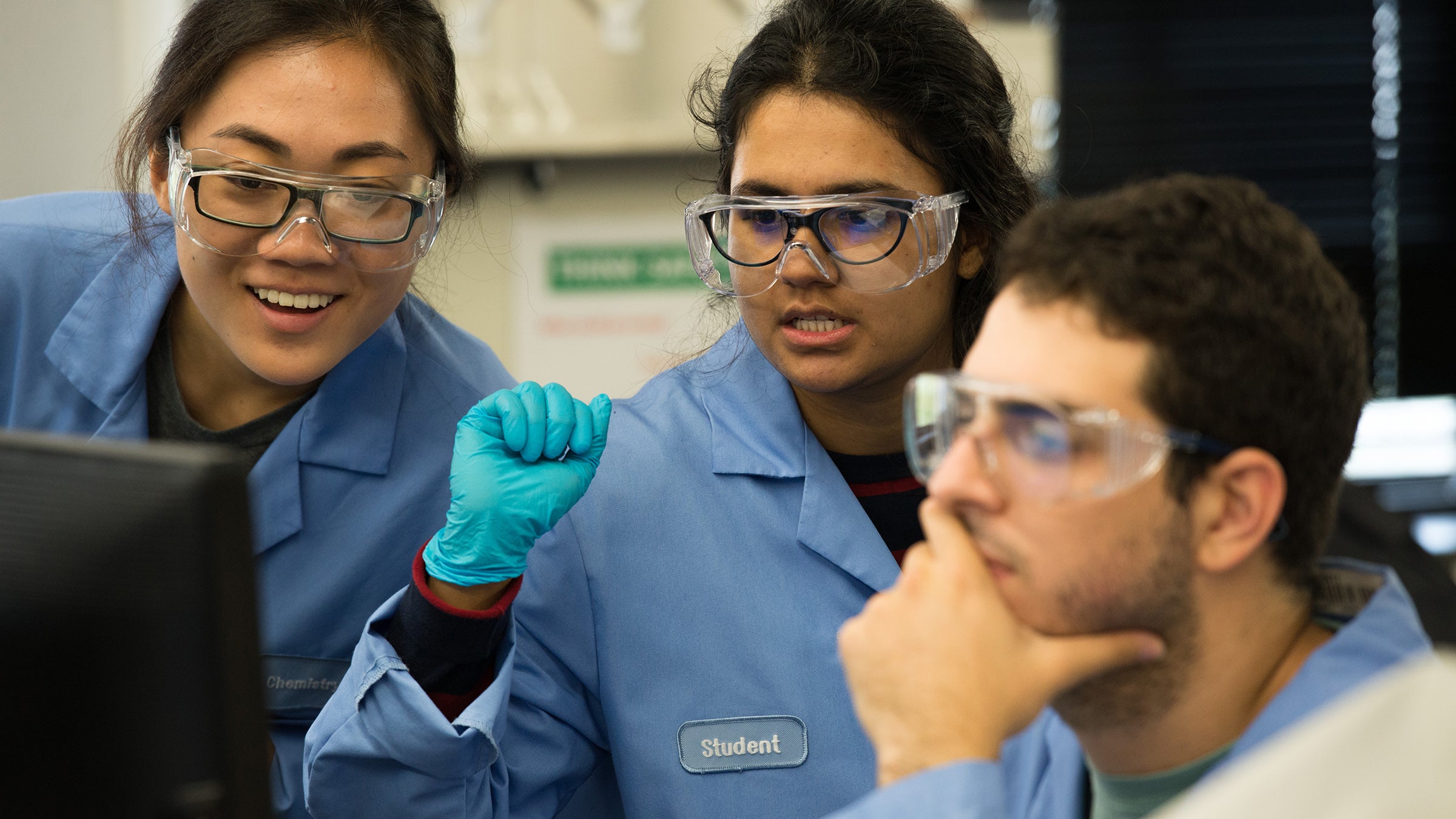 Three students in blue lab coats and goggles gather around a computer screen