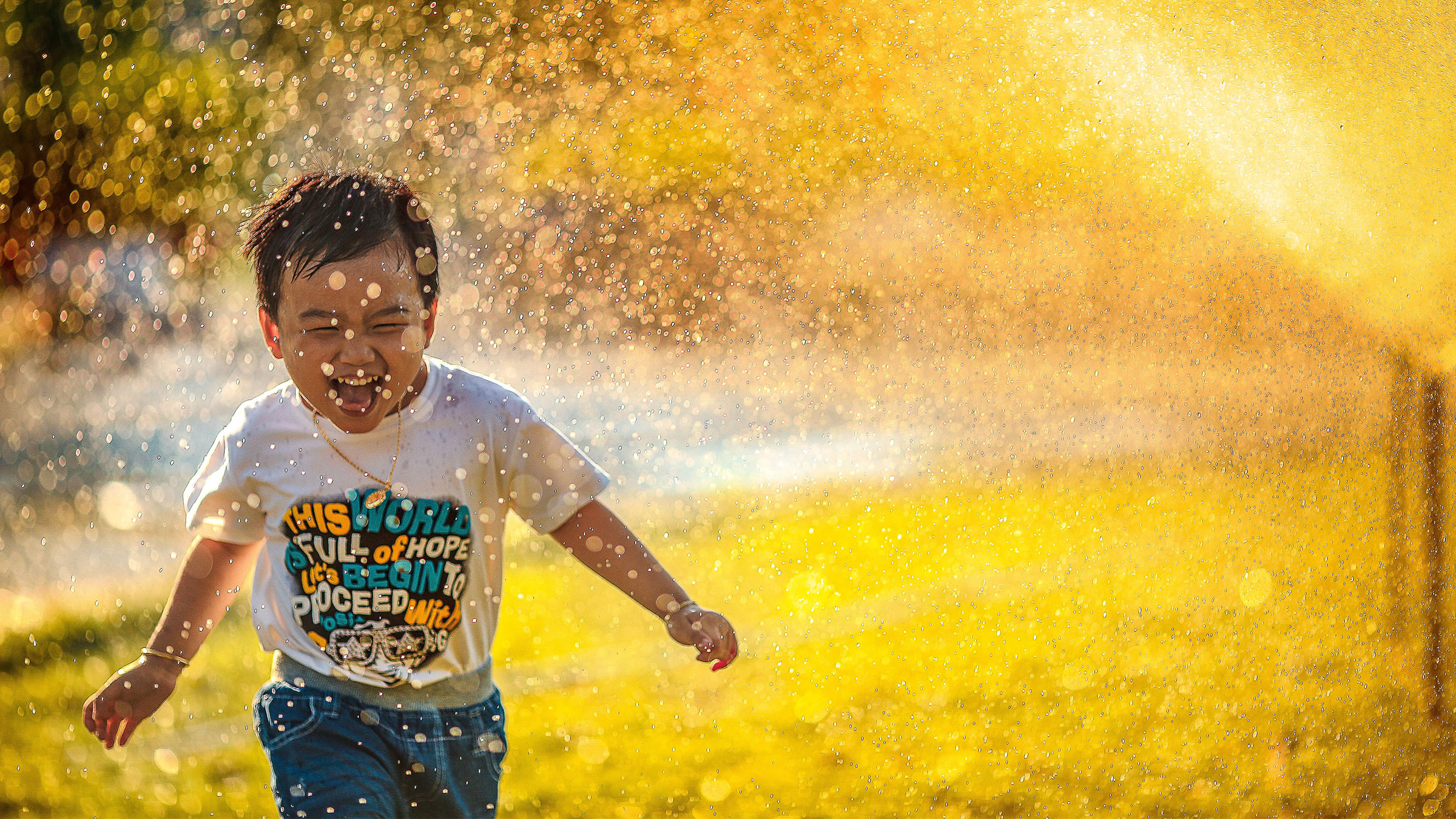 A preschool aged boy plays in a water sprinkler with bright sunlight shining