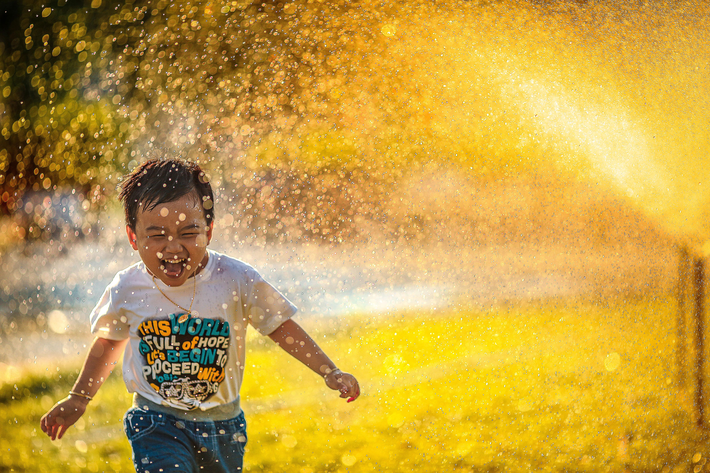 A preschool aged boy plays in a water sprinkler with bright sunlight shining