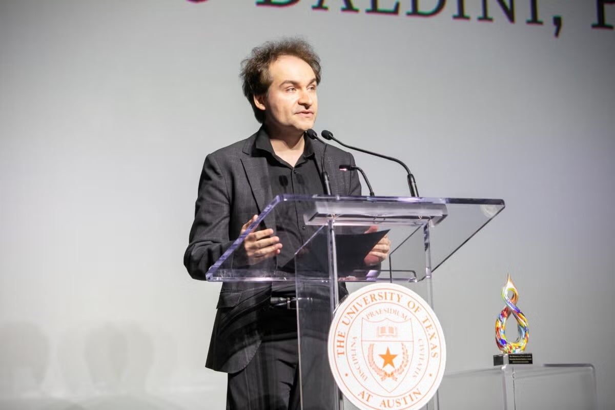 A man stands at a podium addressing an audience. The University seal and an award are on display.