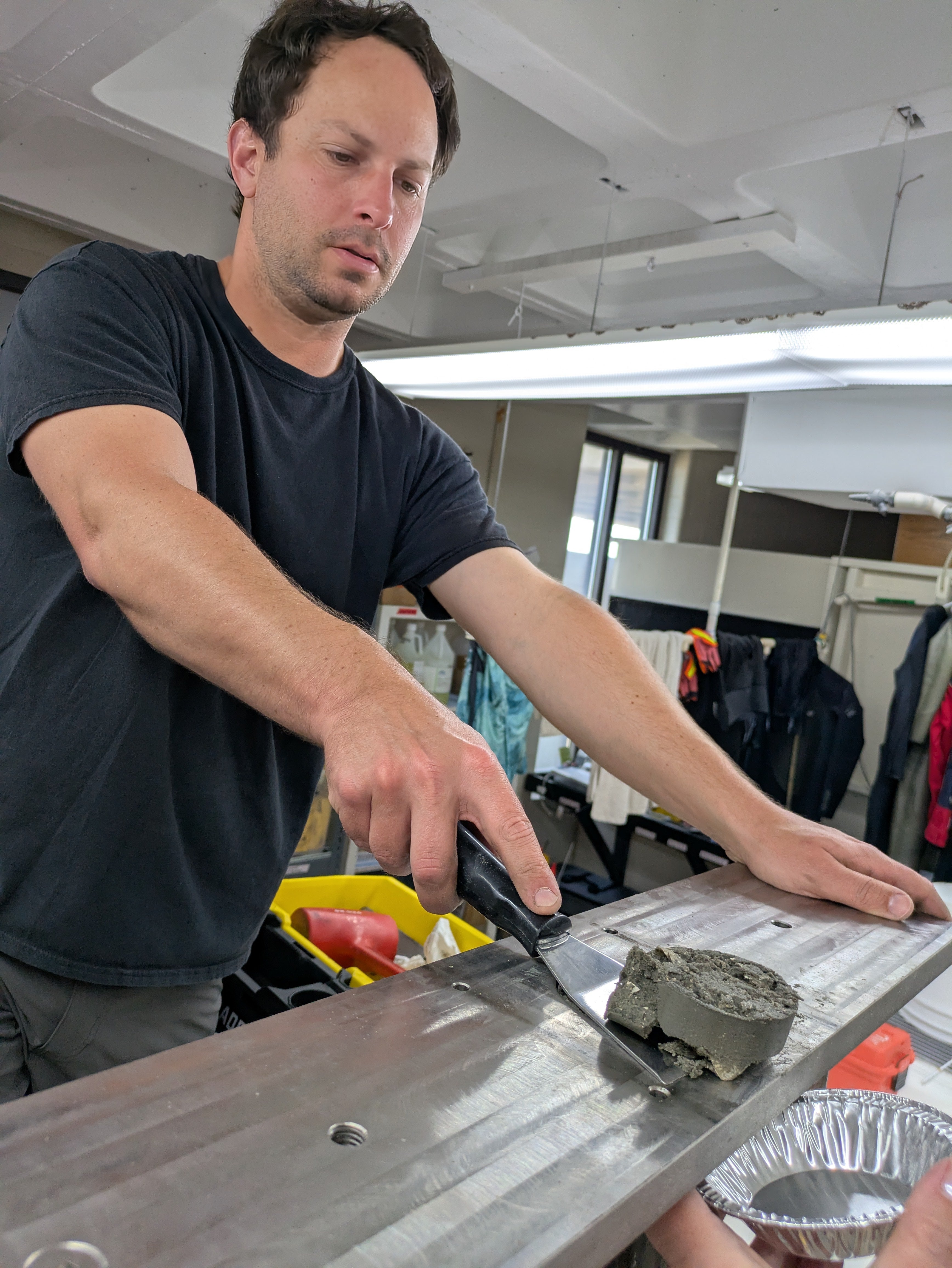 A scientist stands on a ladder and scrapes a grey, hockey puck-sized disc off the top of a metal cross bar.