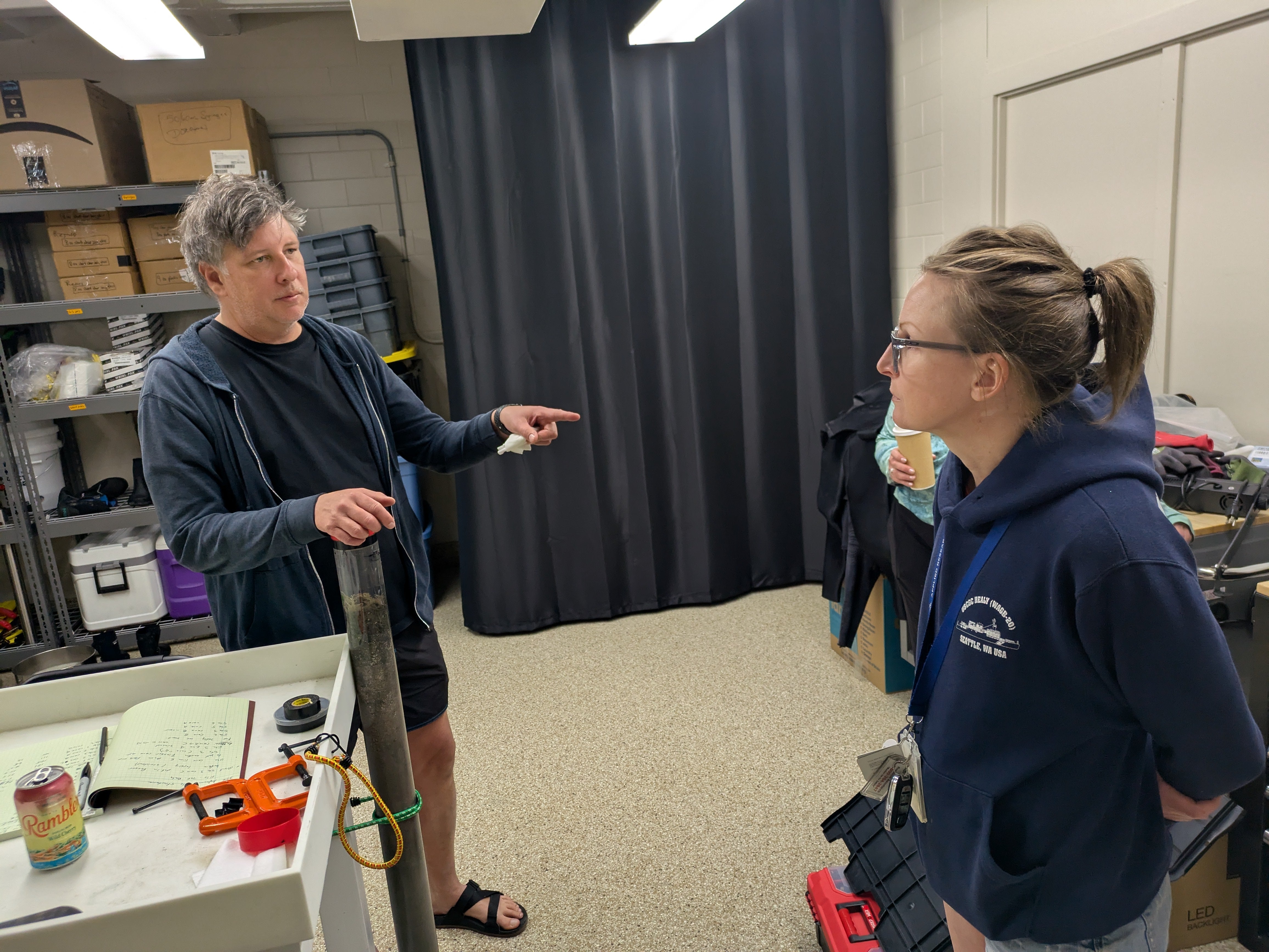 Two scientists talking in a lab. One is holding a four foot long plastic tube containing a sediment core.